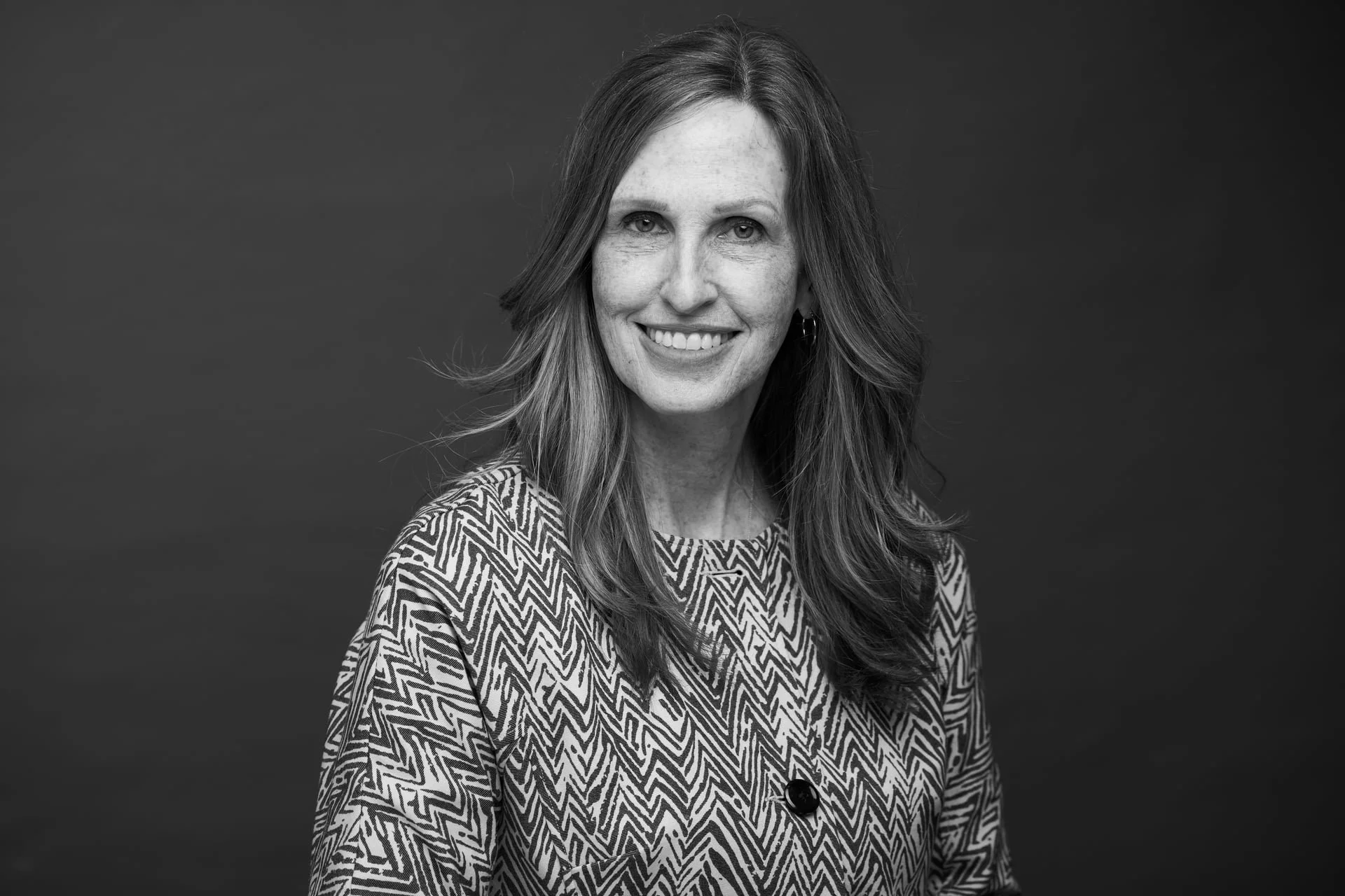 Black and white portrait of a woman smiling, with long wavy hair, wearing a patterned jacket and earrings, against a plain dark background.