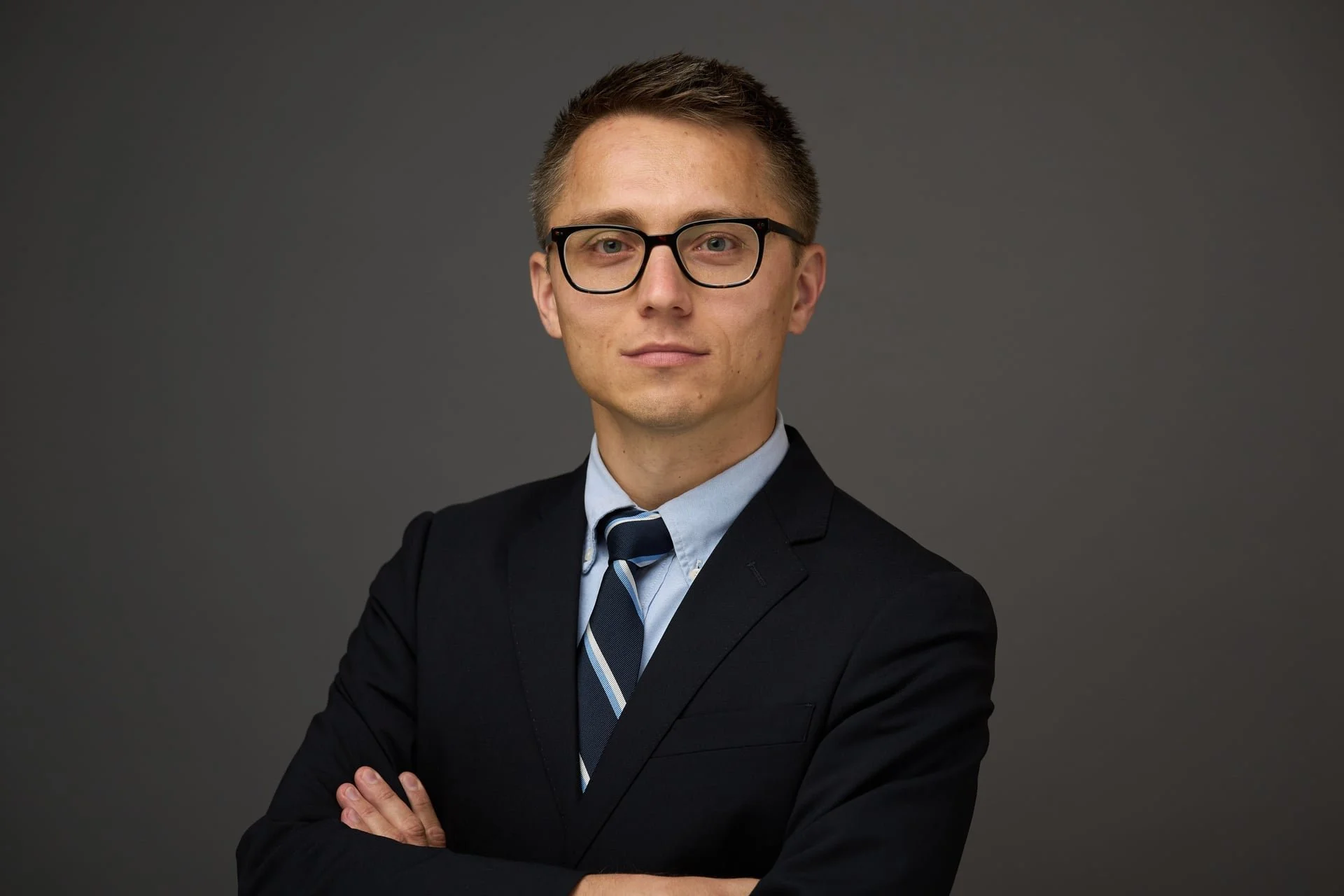 A young man wearing glasses, a dark suit, and a striped tie, with arms crossed, standing against a gray background.