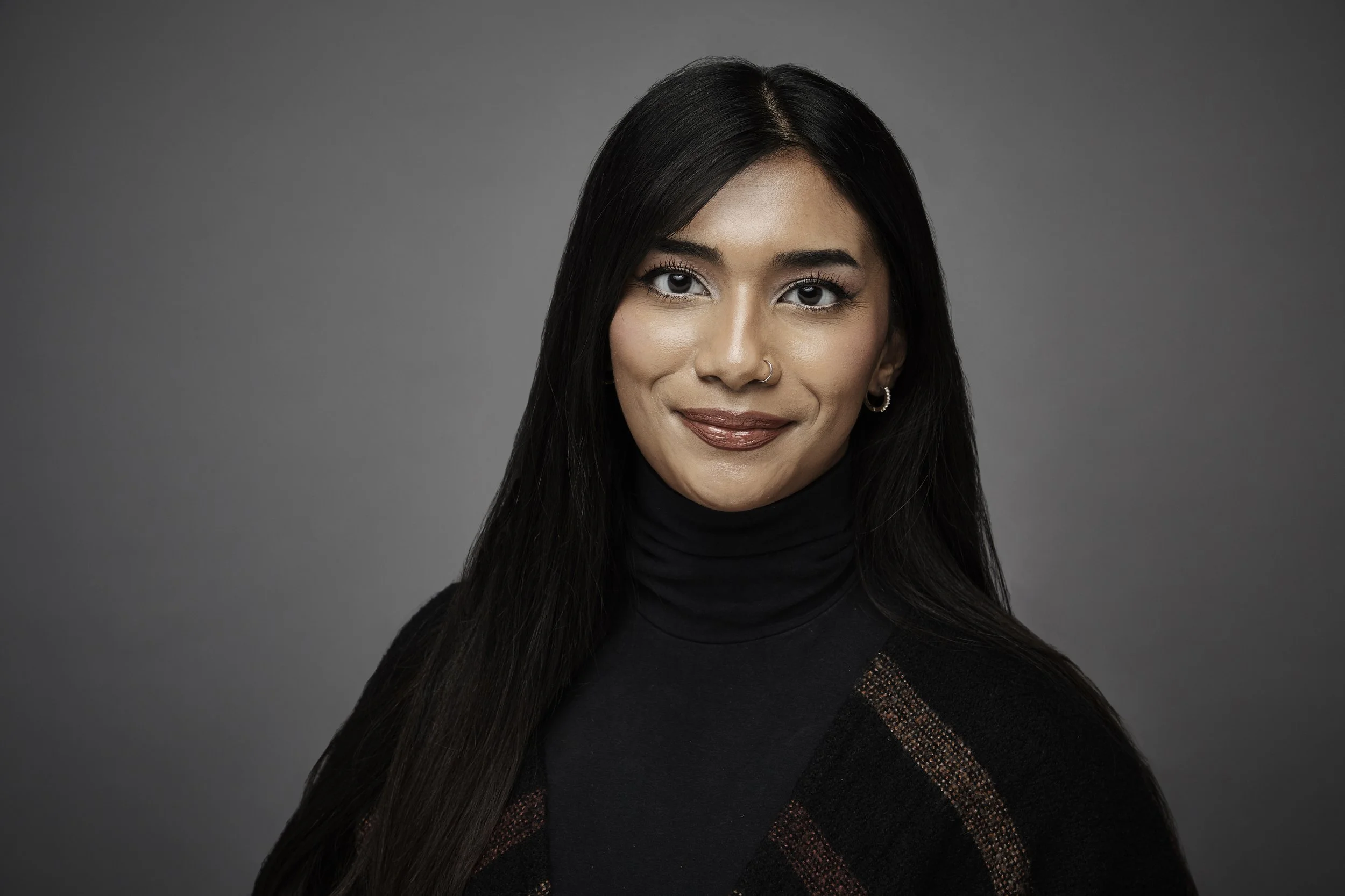 Young woman with long dark hair, hoop earrings, light makeup, wearing a black turtleneck and sweater with a colorful stripe, against a plain gray background.