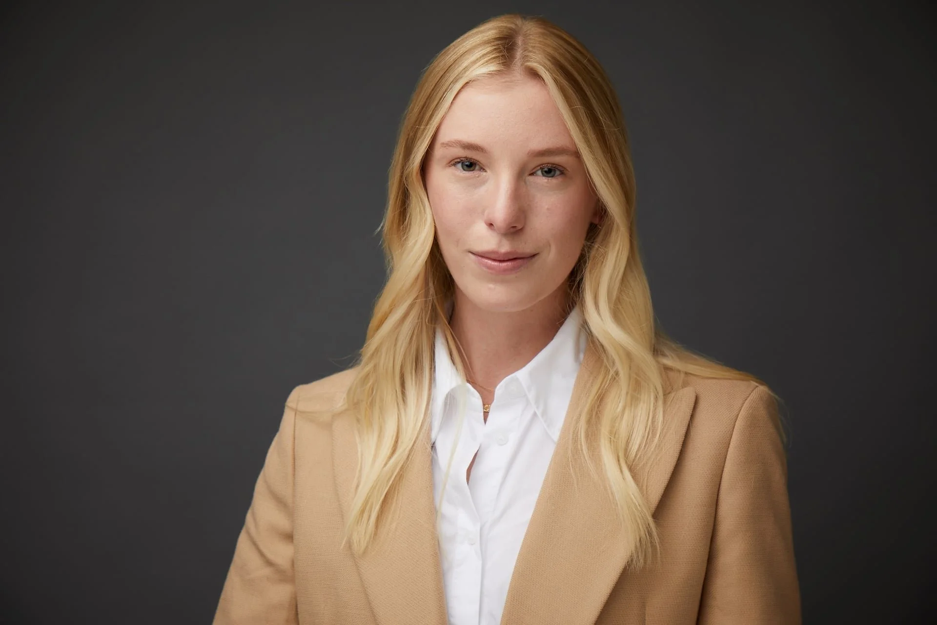 A young woman with long blonde hair, light complexion, dressed in a white shirt and tan blazer, standing against a dark gray background.