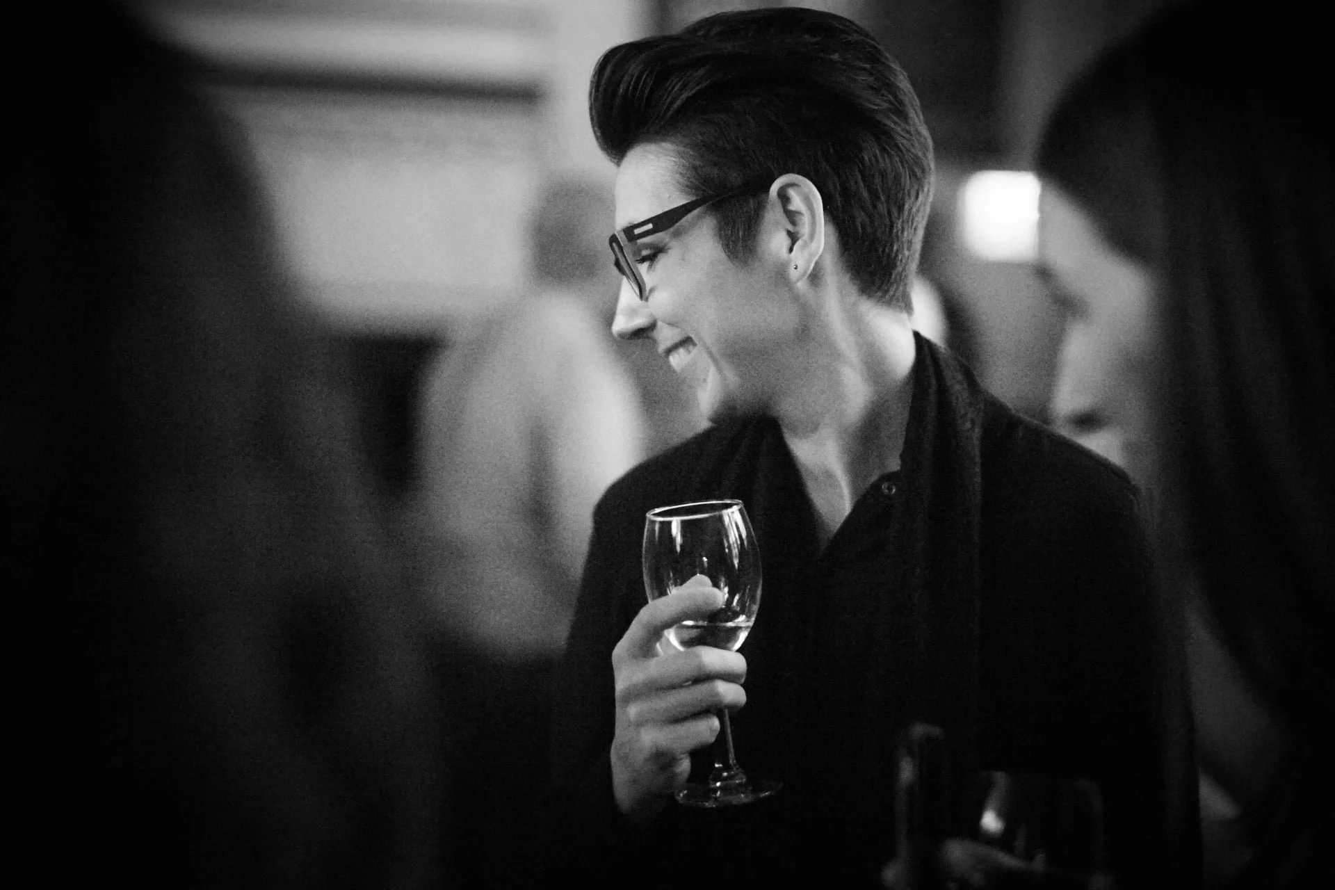 A person with short, styled hair and glasses, smiling while holding a wine glass at a social gathering.