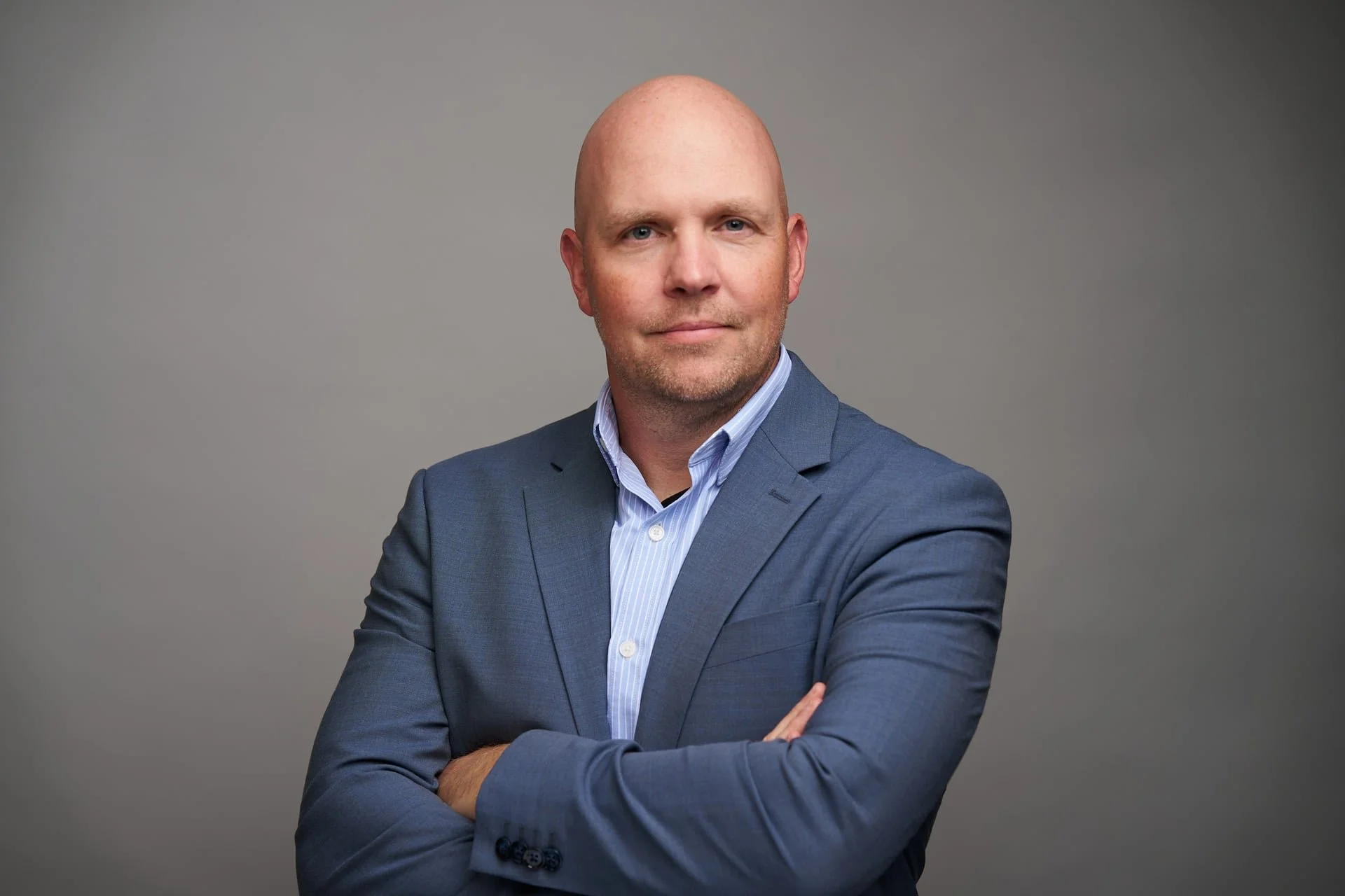 A portrait of a bald man wearing a blue suit and a light blue striped shirt, with arms crossed, against a plain gray background.