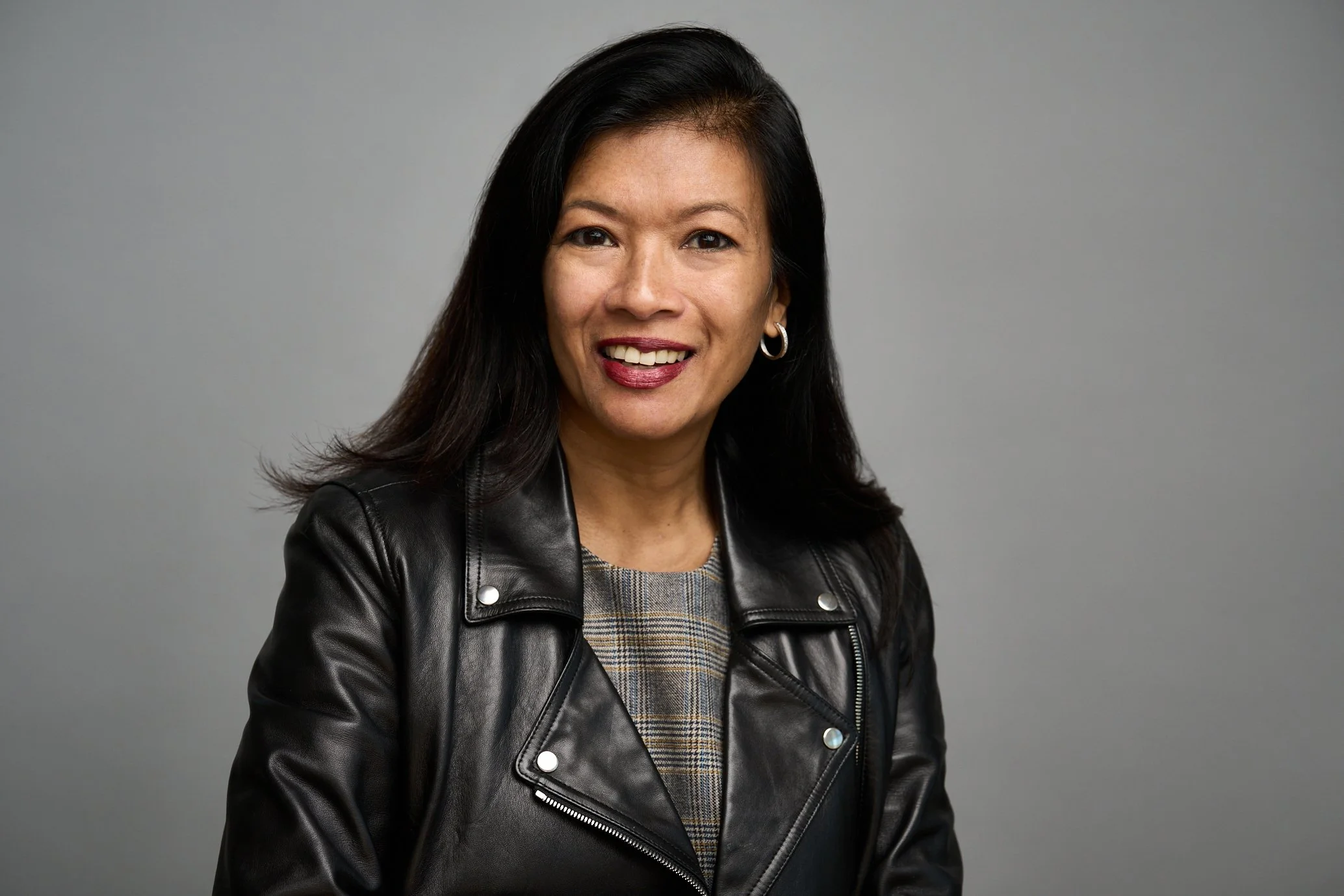 A woman with long dark hair, wearing hoop earrings, a plaid shirt, and a black leather jacket, smiling against a gray background.