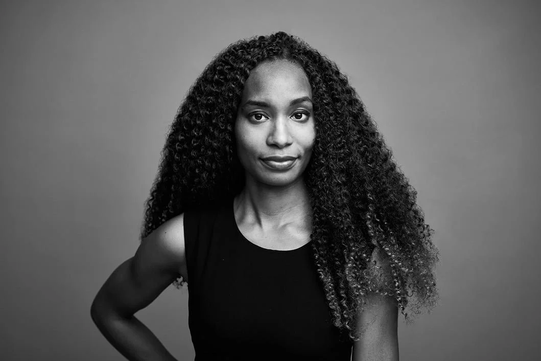 Black and white portrait of a woman with long curly hair wearing a sleeveless top, standing against a plain background.