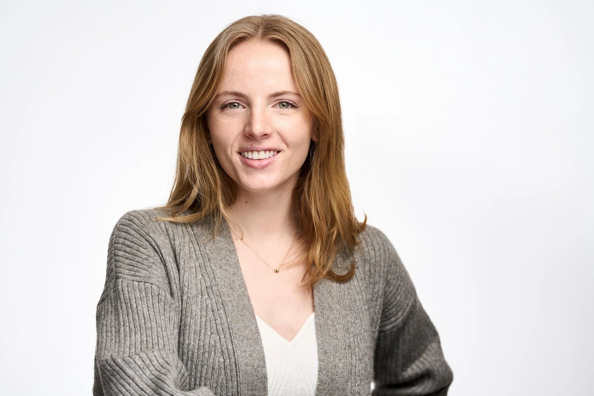 Portrait of a young woman with red hair, green eyes, smiling, wearing a beige sweater and a white top, against a plain white background.