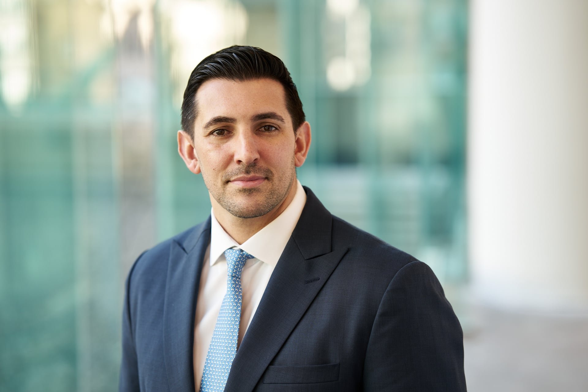 A portrait of a young man in a navy blue suit, white shirt, and light blue tie, standing in front of a blurred modern office building with glass panels.