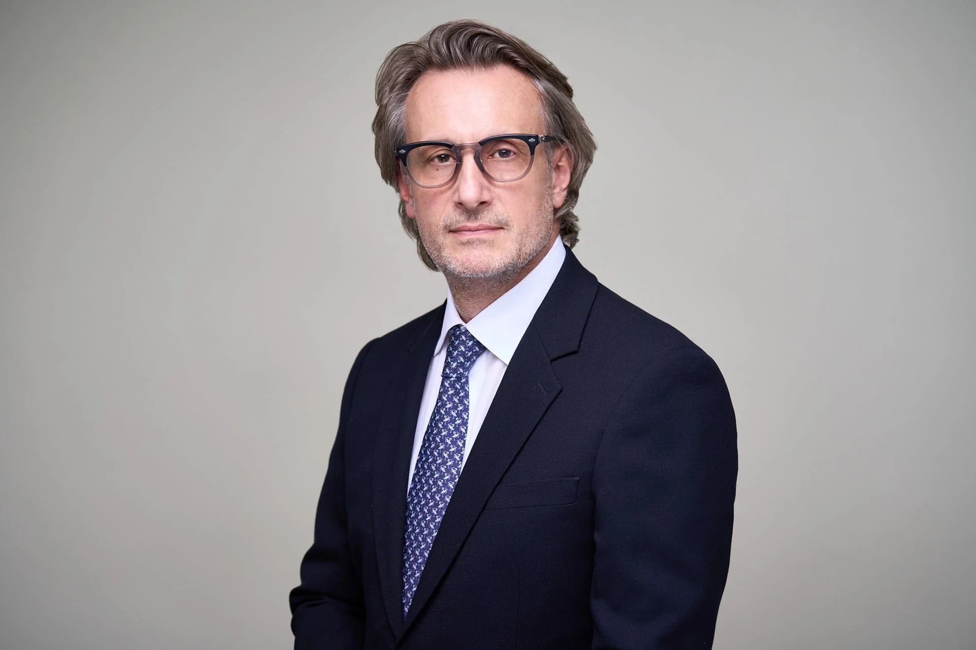 A middle-aged man with gray hair and glasses, wearing a navy suit, white shirt, and patterned tie, posing against a plain gray background.