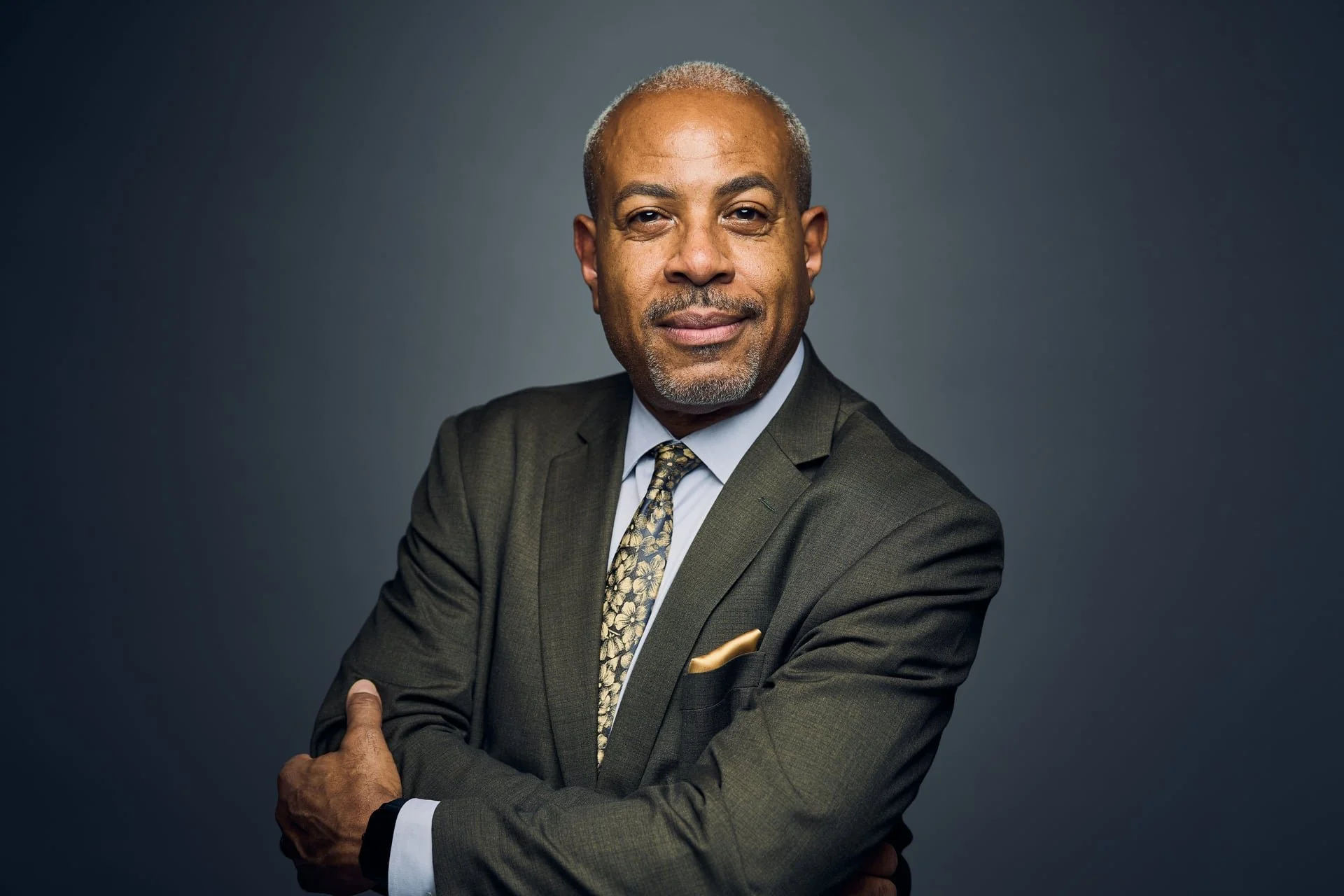 A confident middle-aged African American man dressed in a gray suit, light blue shirt, and patterned tie, with arms crossed, standing against a dark gray background.