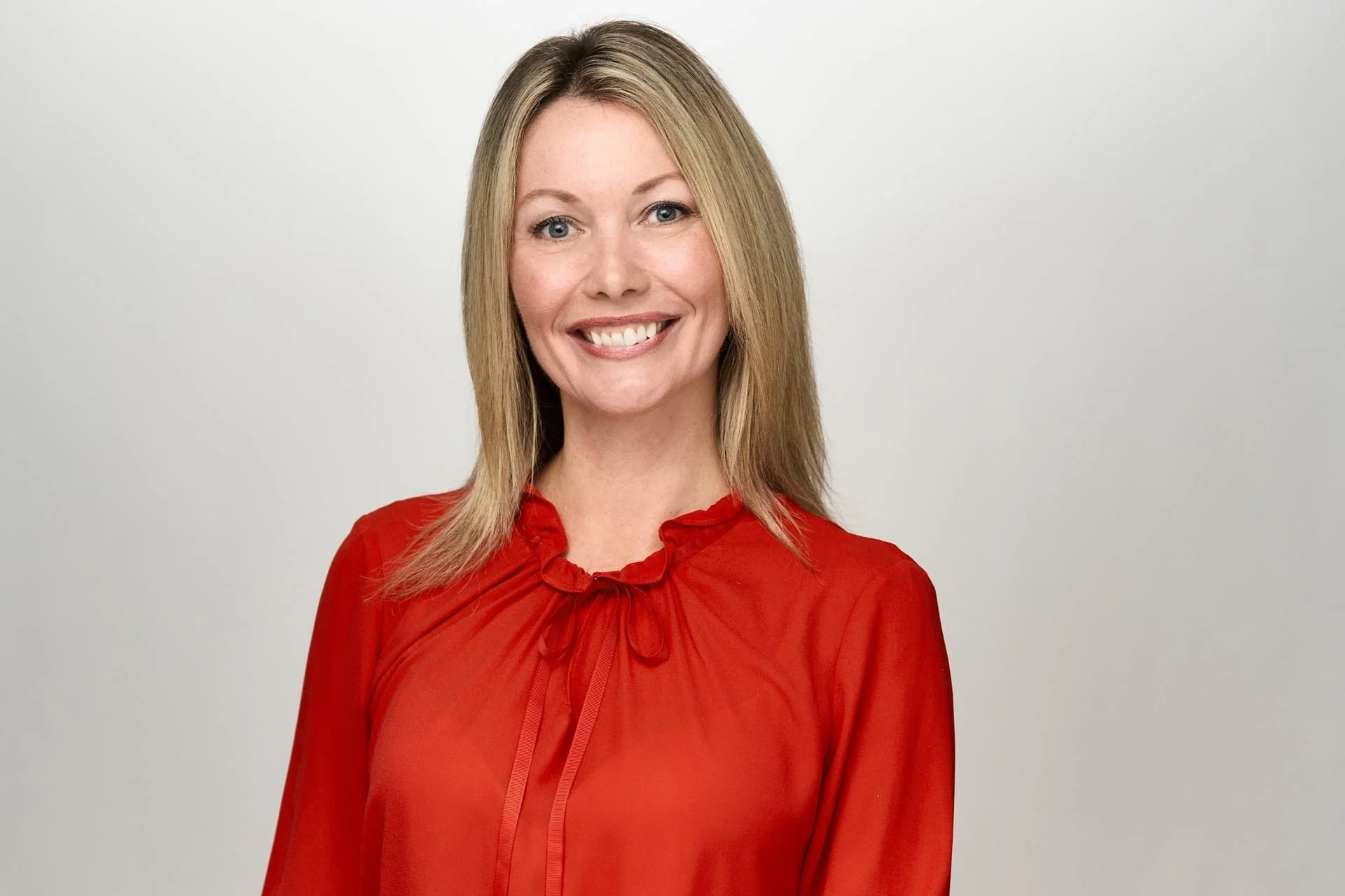 A woman with blonde hair, wearing a red blouse with a ruffled collar and a ribbon tied at the neck, smiling against a plain light gray background.
