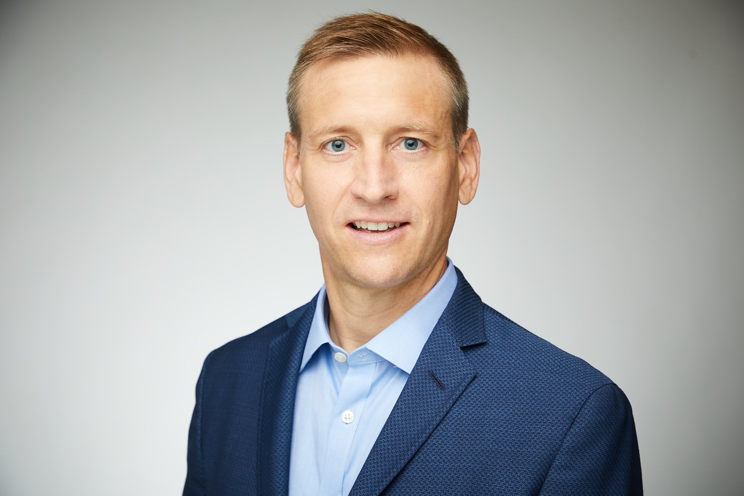 Headshot of a man with short light brown hair, blue eyes, wearing a blue suit and a light blue dress shirt, against a plain gray background.
