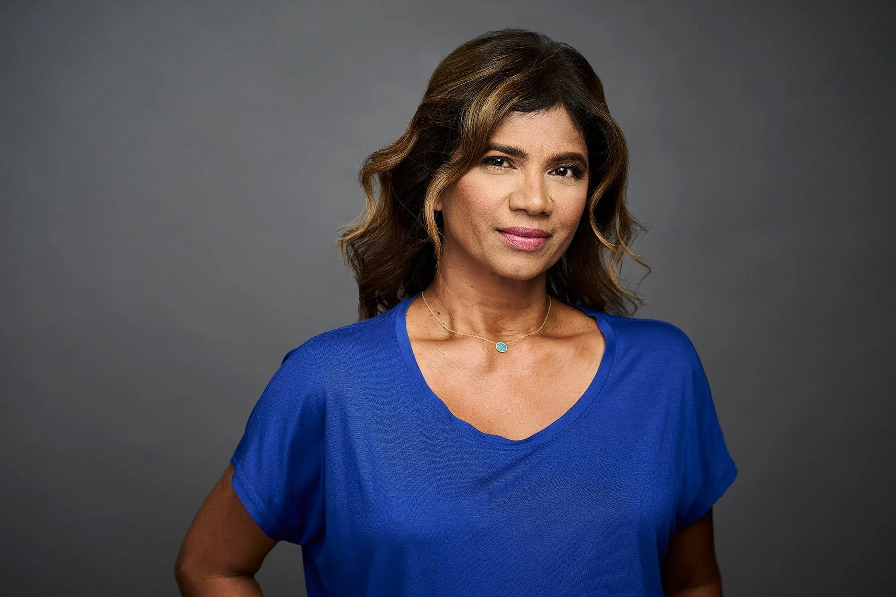 A woman with medium-length brown hair styled in loose waves, wearing a blue top and a delicate necklace with a blue pendant, standing against a plain gray background.