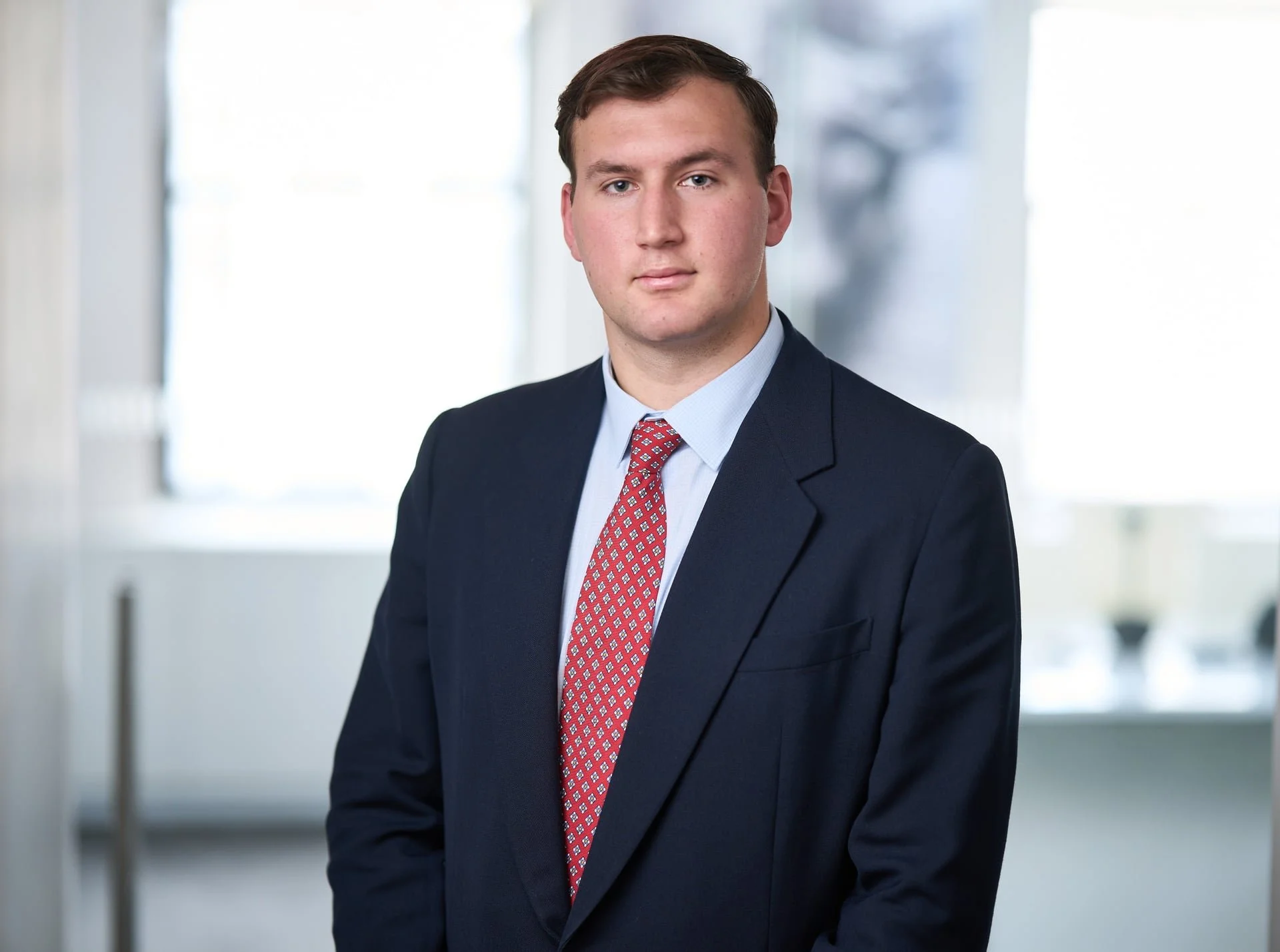 Professional man in a dark suit and red patterned tie standing in a modern office with large windows.