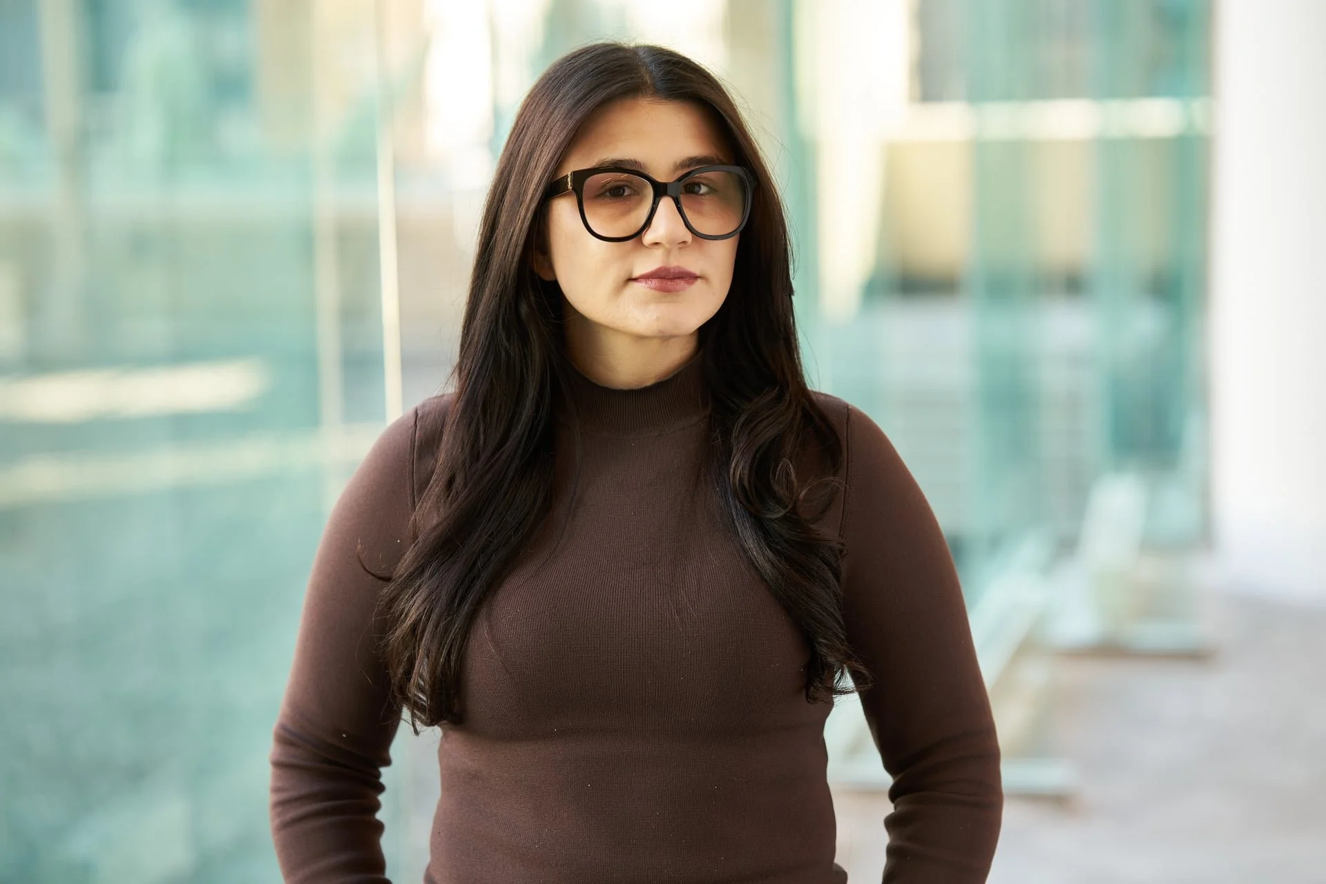 A woman with long dark hair wearing glasses and a brown turtleneck sweater, standing in front of a glass building.