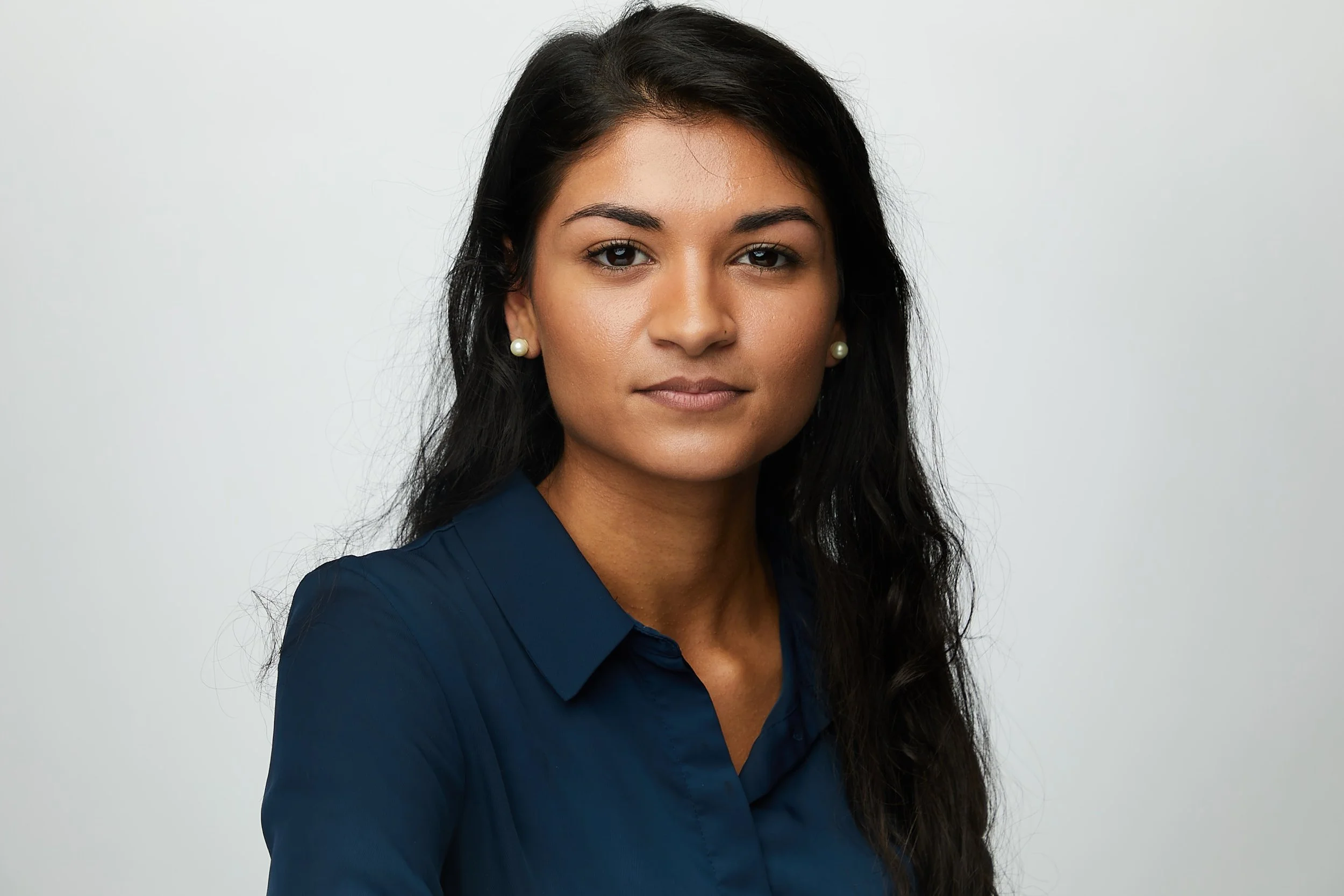 A woman with long dark hair, wearing pearl earrings and a navy blue shirt, poses against a plain light-colored background.
