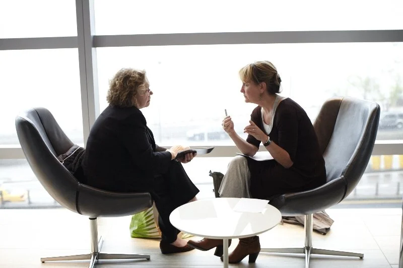 Two women having a discussion in a modern lounge area with large windows, one holding a tablet and the other gesturing with a pen.