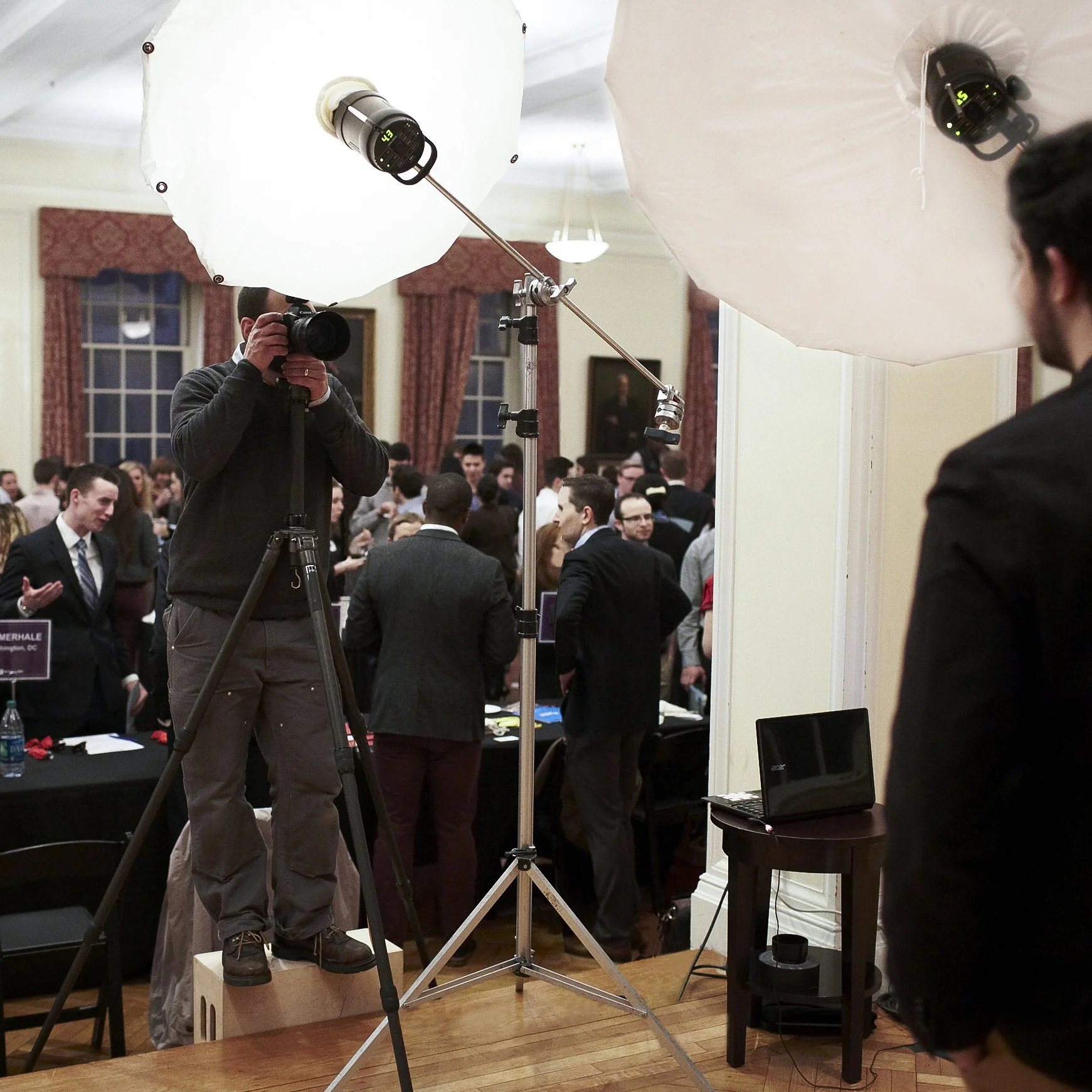 Photographer taking a picture at a formal event with people in suits, set in an elegant room with large windows, red curtains, and a portrait painting on the wall.