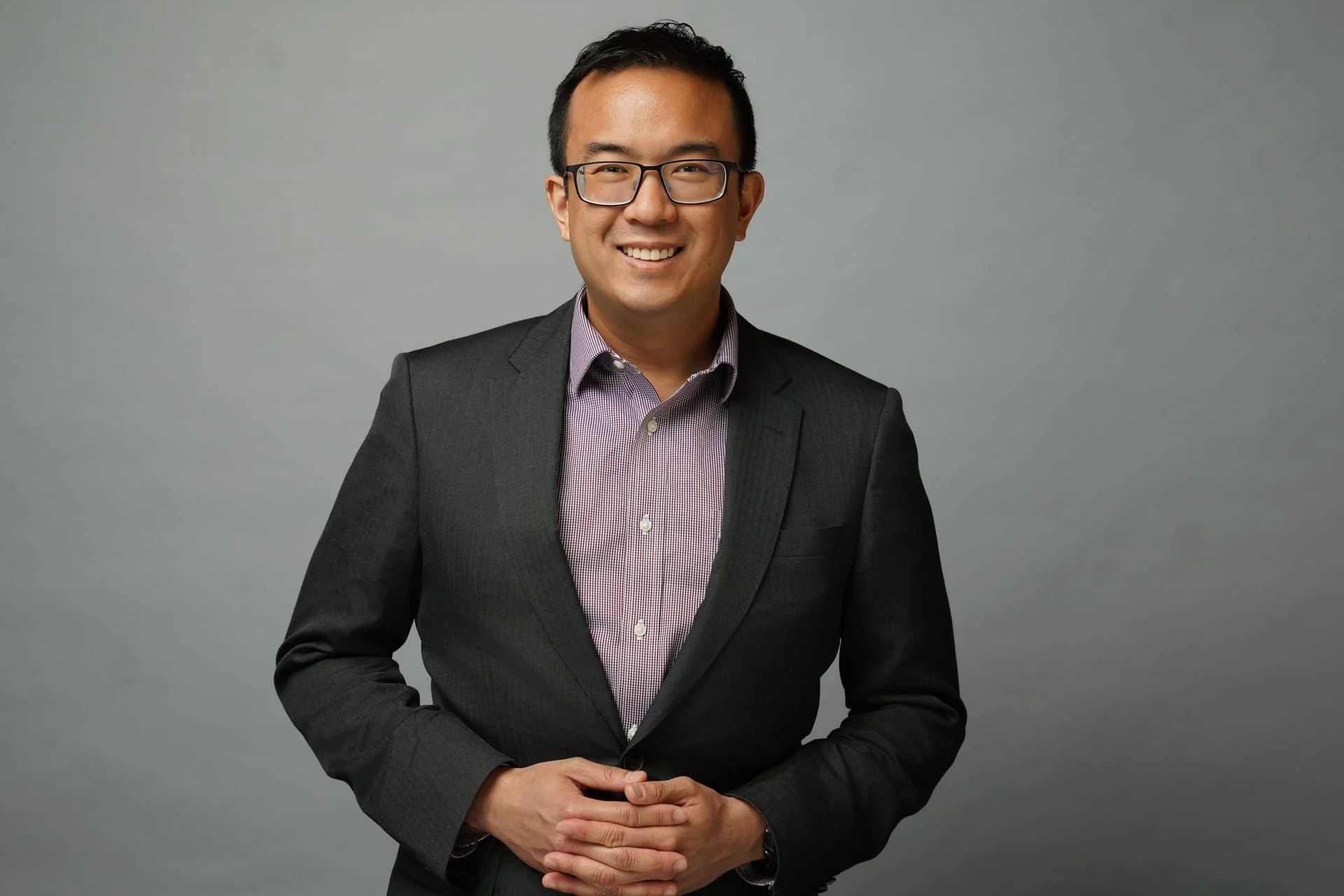 A man in a dark gray suit and glasses standing against a gray background, smiling with hands clasped in front.