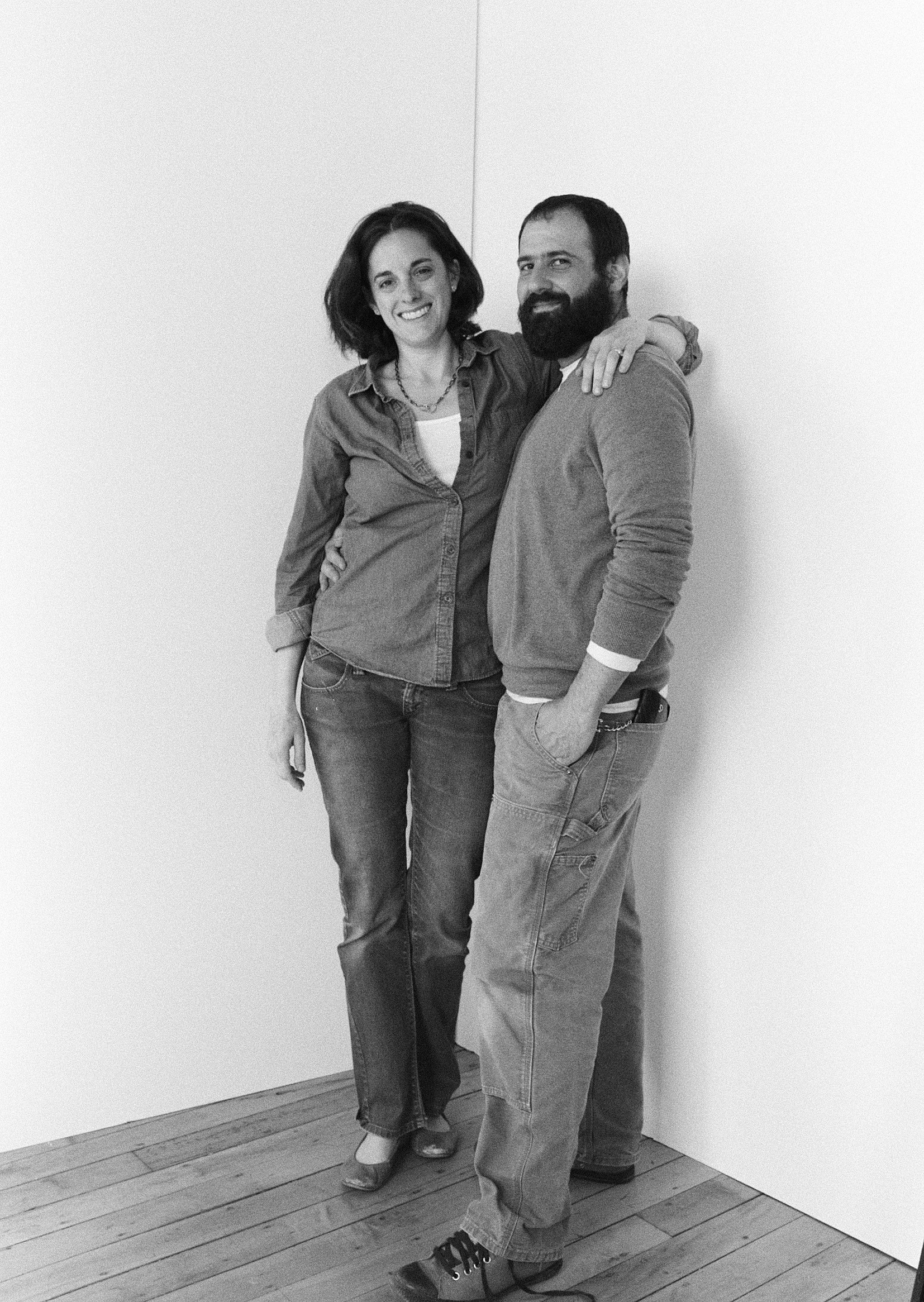 A smiling woman with shoulder-length hair and a man with a beard, both dressed casually and standing close together against a plain wall, with the woman's arm around the man's shoulders.