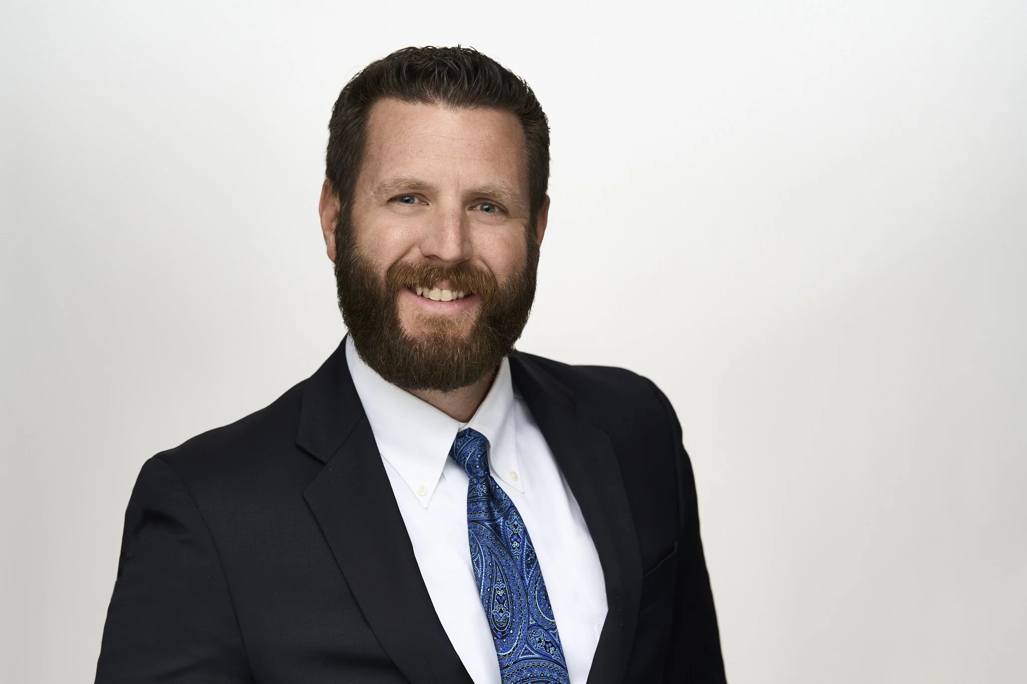 A man with a beard and short hair wearing a black suit, white shirt, and a blue patterned tie, smiling against a plain white background.