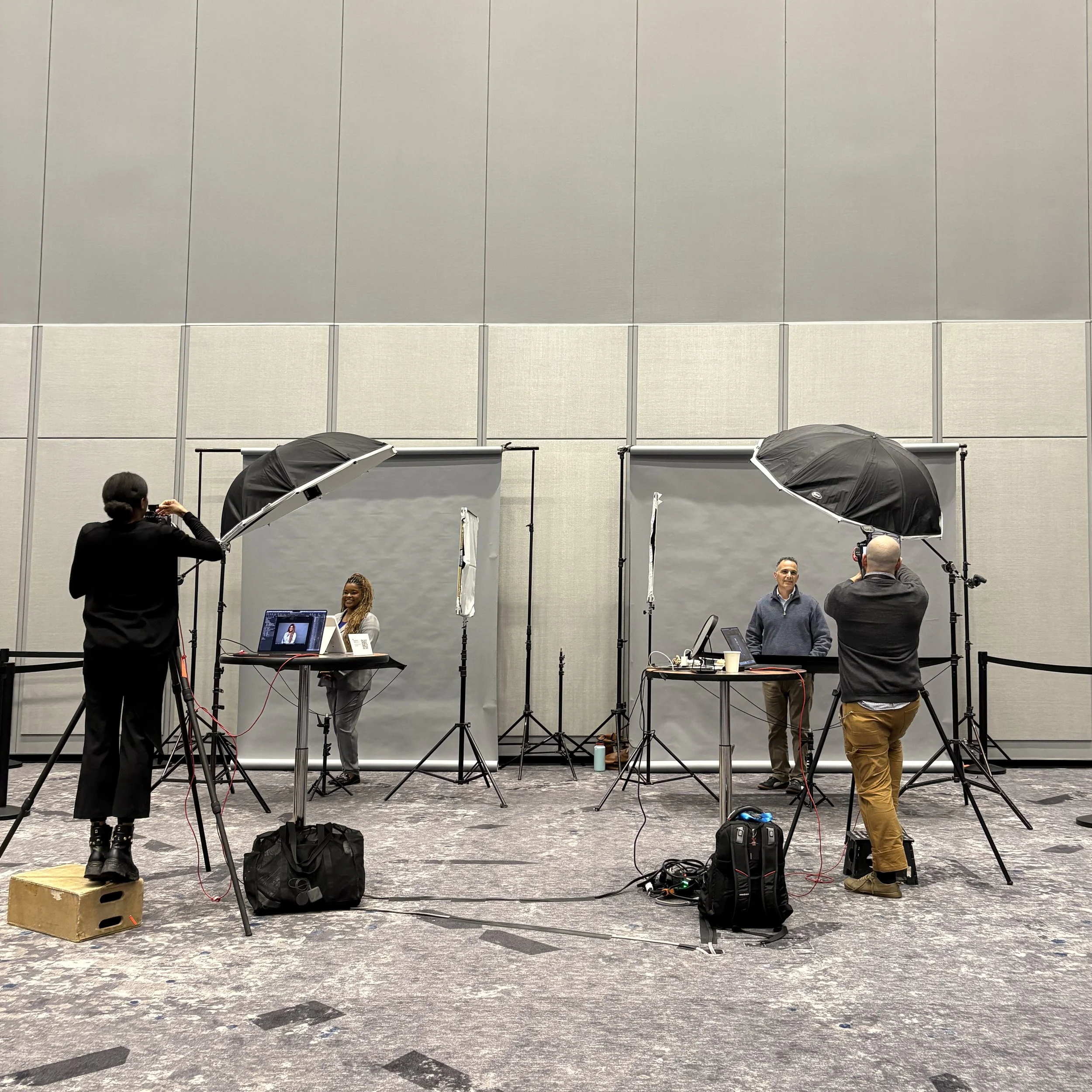 Behind the scenes at a photography or video shoot, with lighting equipment, umbrellas, and people preparing and working.