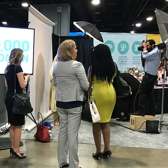 Women at a trade show or conference, some waiting in line and one taking a photo with a professional camera.