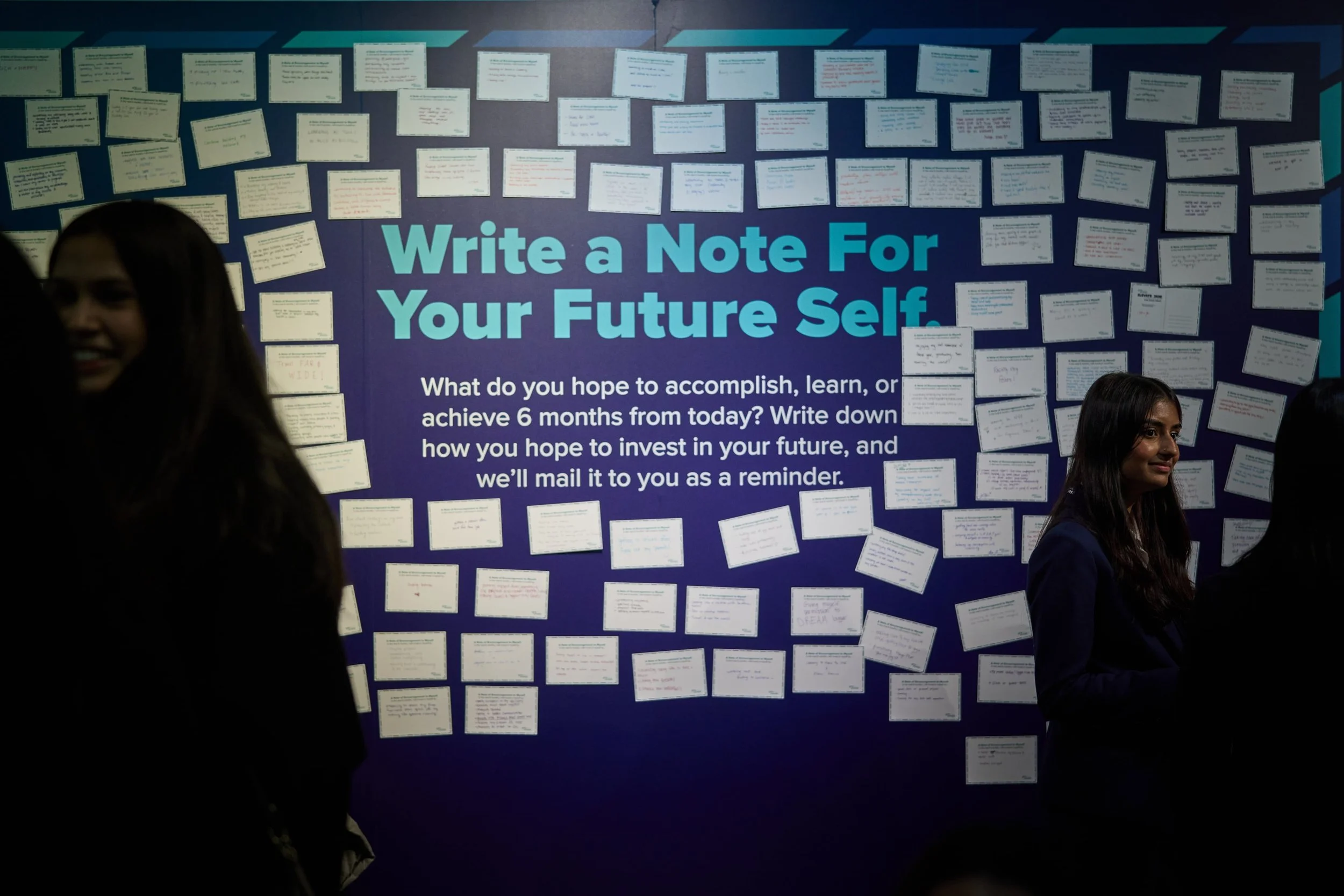 A bulletin board with the message 'Write a Note For Your Future Self' in large blue text. The board is filled with numerous handwritten notes on small white cards arranged in rows and columns. Several people are standing in front of the board, some p
