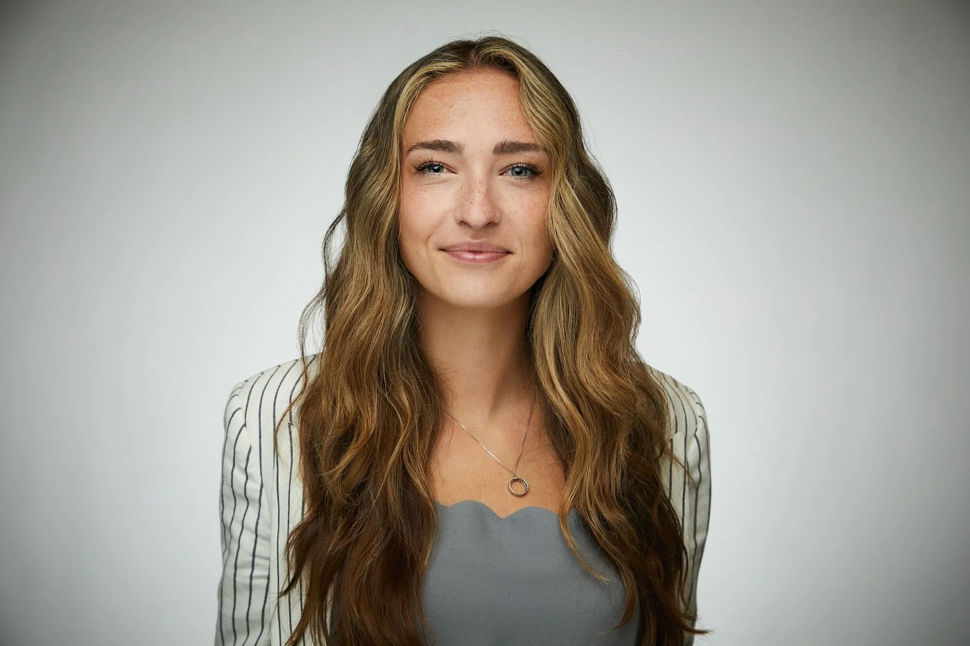 A young woman with long, wavy light brown hair, fair skin with freckles, and blue eyes. She is wearing a gray top with a circular necklace and a white blazer with black pinstripes. She is smiling softly and winking, standing against a plain light gra