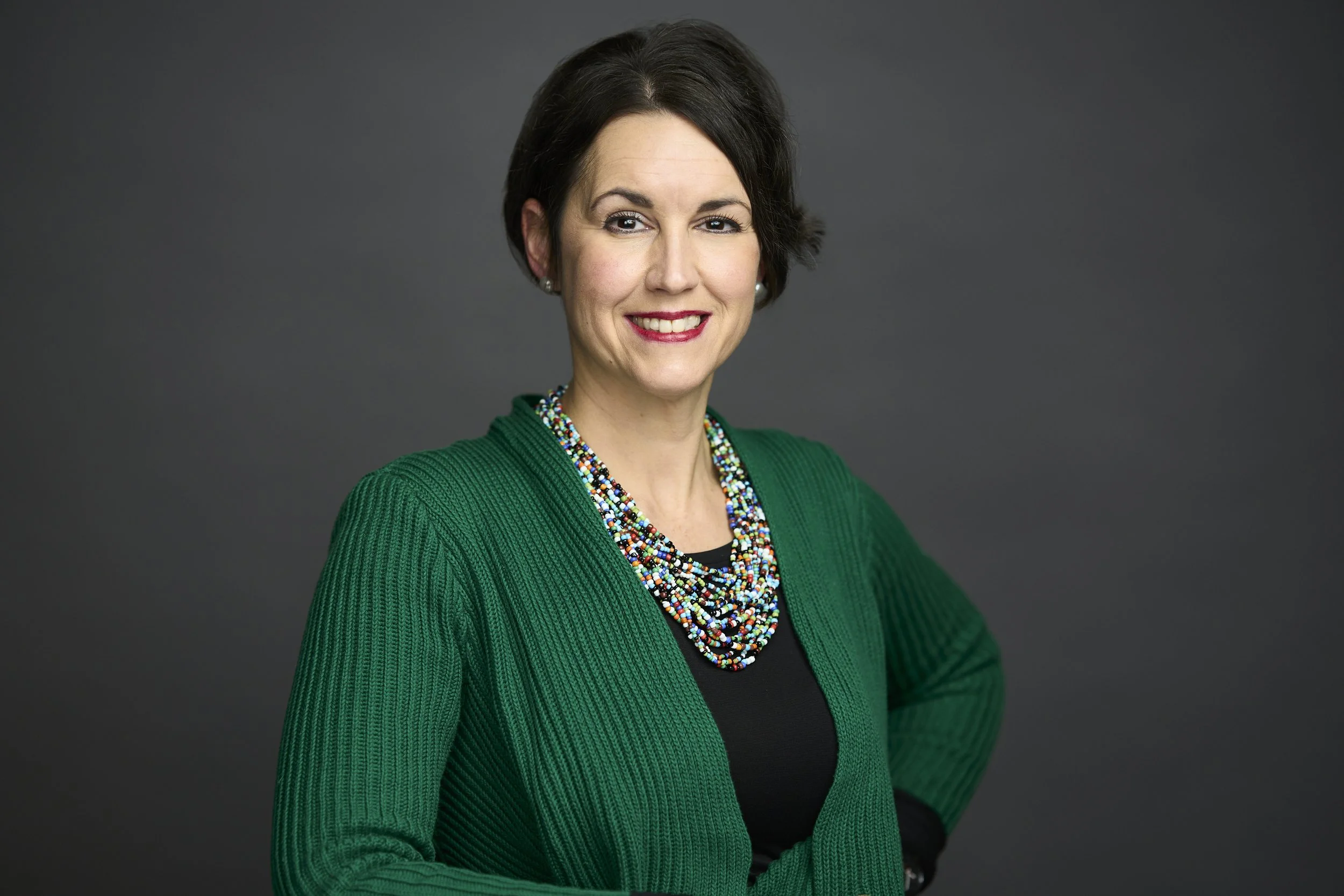 Portrait of a smiling woman with short dark hair, wearing a green sweater and colorful beaded necklaces, against a dark gray background.
