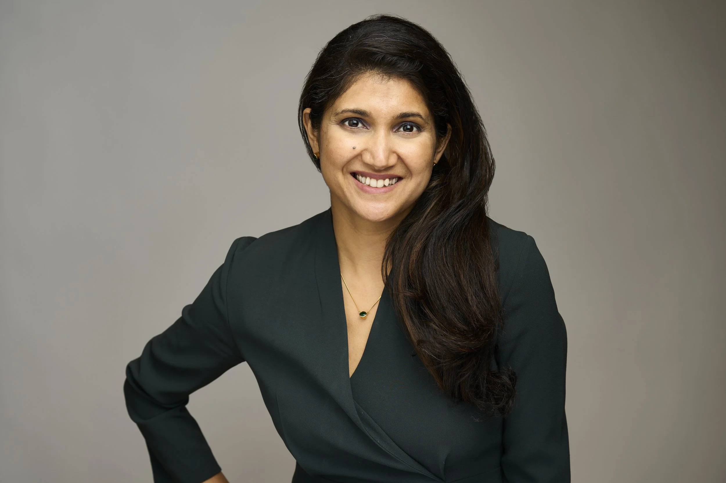A woman with long dark hair, wearing a black blazer with a V-neck shirt, gold earrings, and a pendant necklace, smiling and posing against a plain gray background.