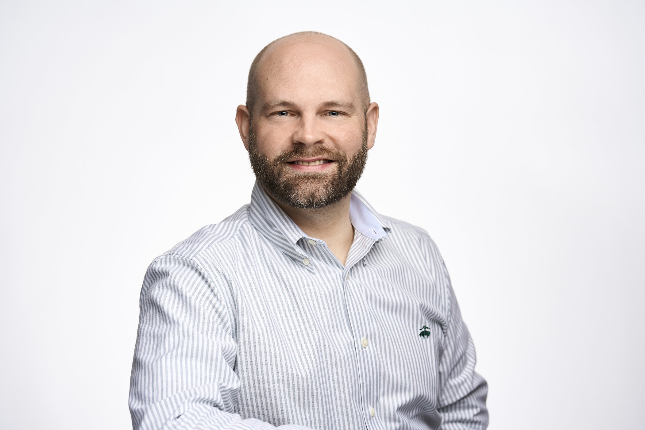 A man with a beard and a shaved head, wearing a light blue striped collared shirt, smiling at the camera against a plain white background.