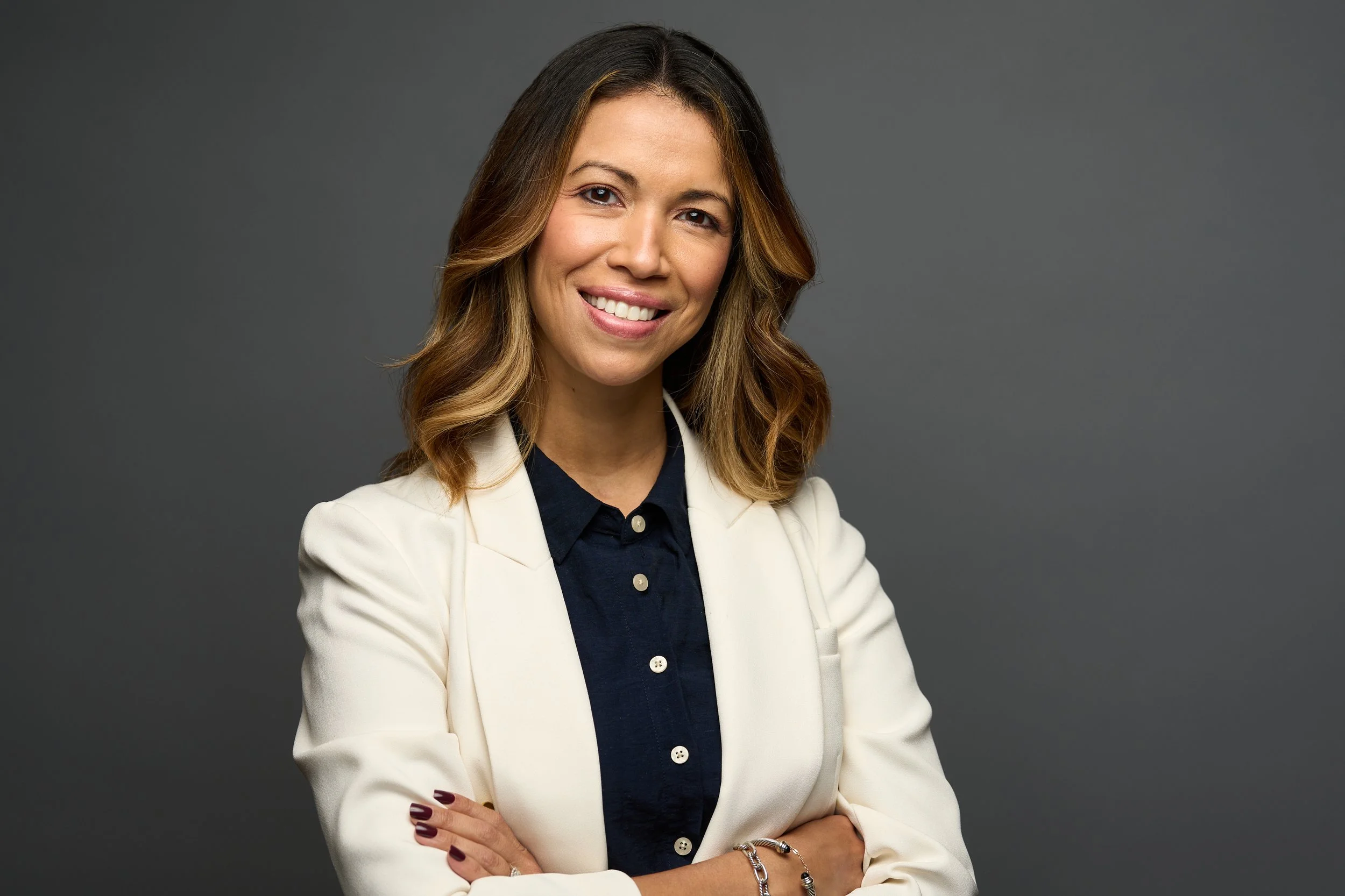A woman with shoulder-length brown hair and highlights, wearing a white blazer over a dark shirt, smiling with crossed arms, against a plain gray background.