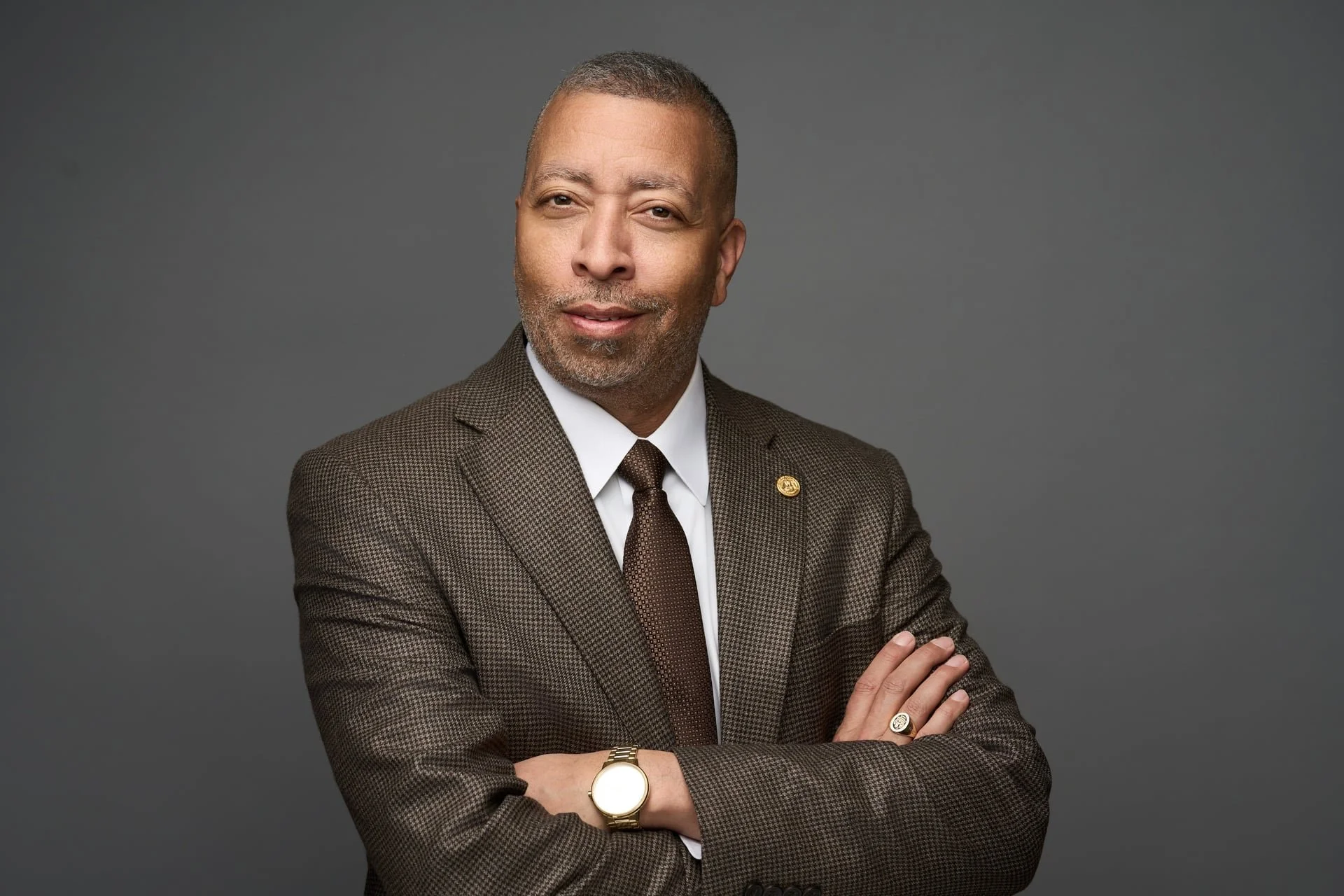 A middle-aged man wearing a brown checked suit, white shirt, and brown tie, with folded arms, looking at the camera with a slight smile, against a gray background.