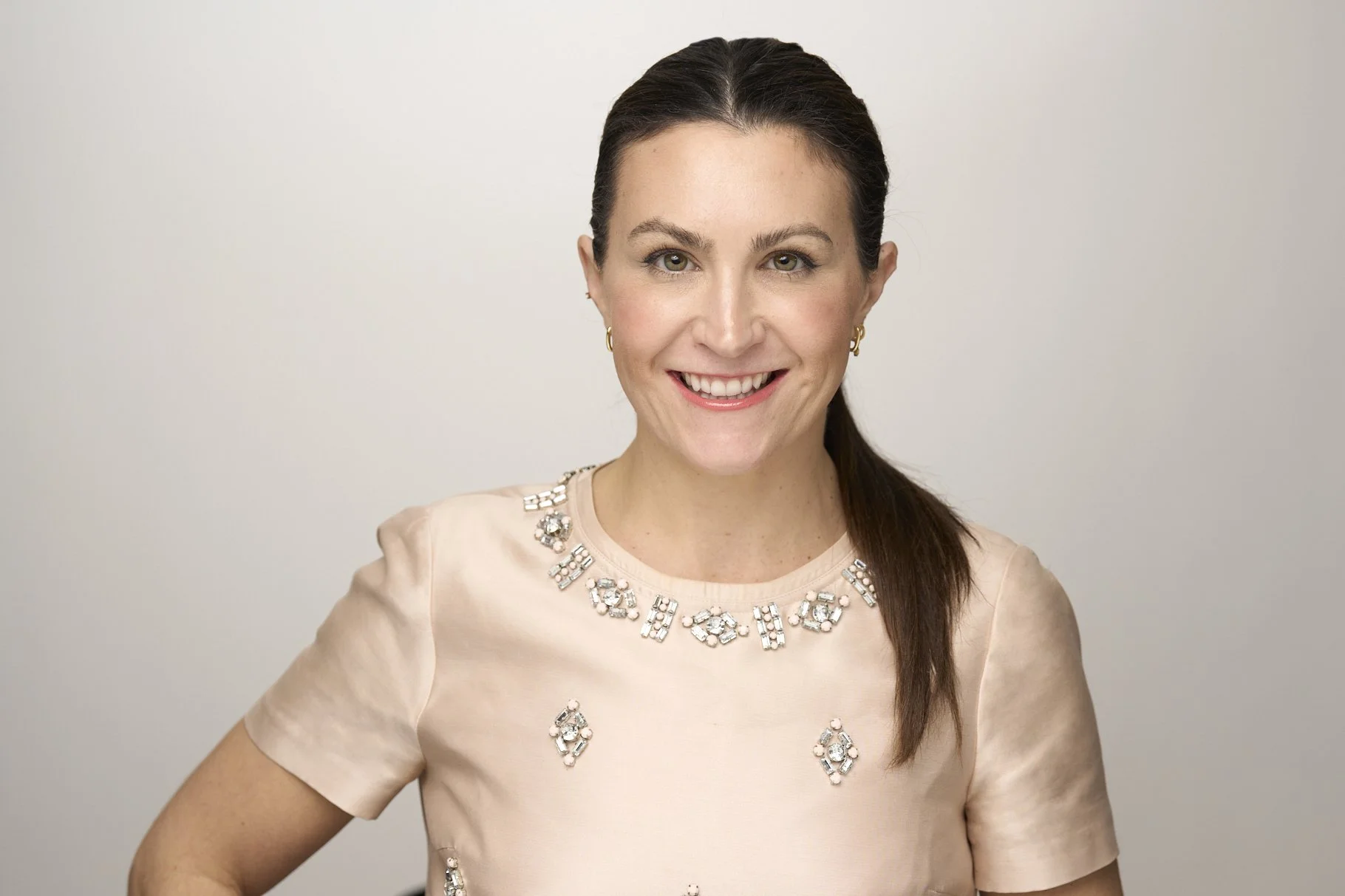 A woman with dark brown hair styled in a ponytail, smiling and wearing a light-colored dress with decorative embellishments around the neckline.