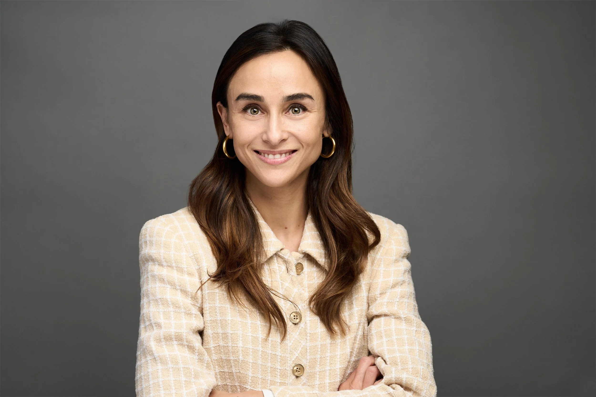 A young woman with long brown hair, light skin, wearing a beige plaid jacket with buttons and gold hoop earrings, smiling and standing against a dark gray background.