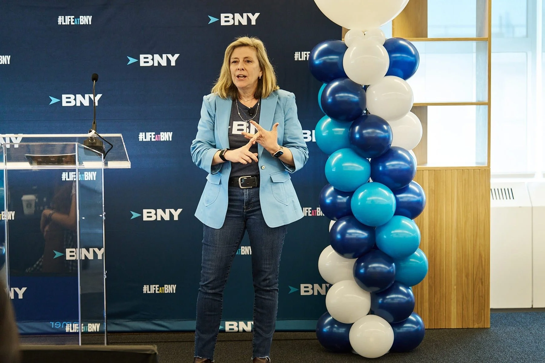 A woman speaking at a BNY event stands near a tall column of blue, white, and navy balloons on the right. She is wearing a light blue blazer, dark jeans, and a black T-shirt with the BNY logo, gesturing with her hands. A backdrop behind her features 