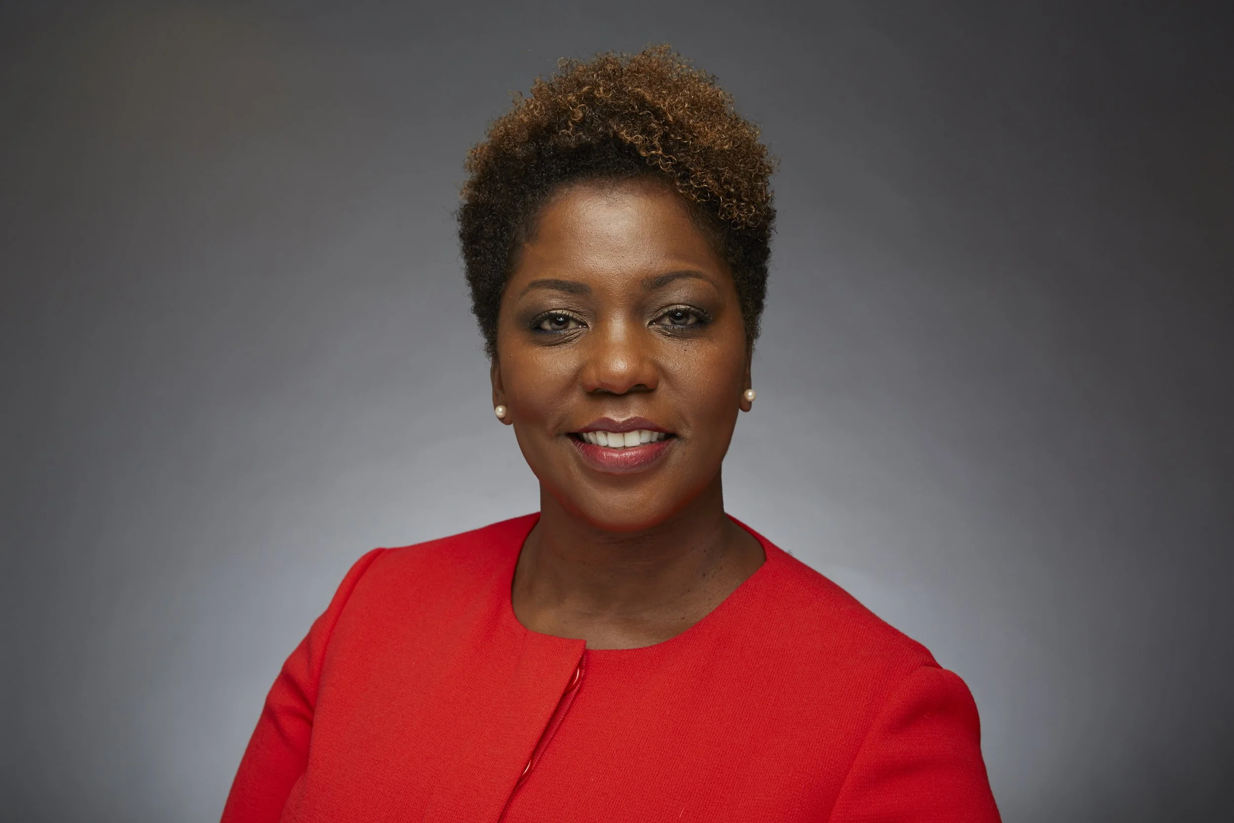 A confident woman with short, curly hair, wearing a red blazer, smiling, against a neutral gray background.