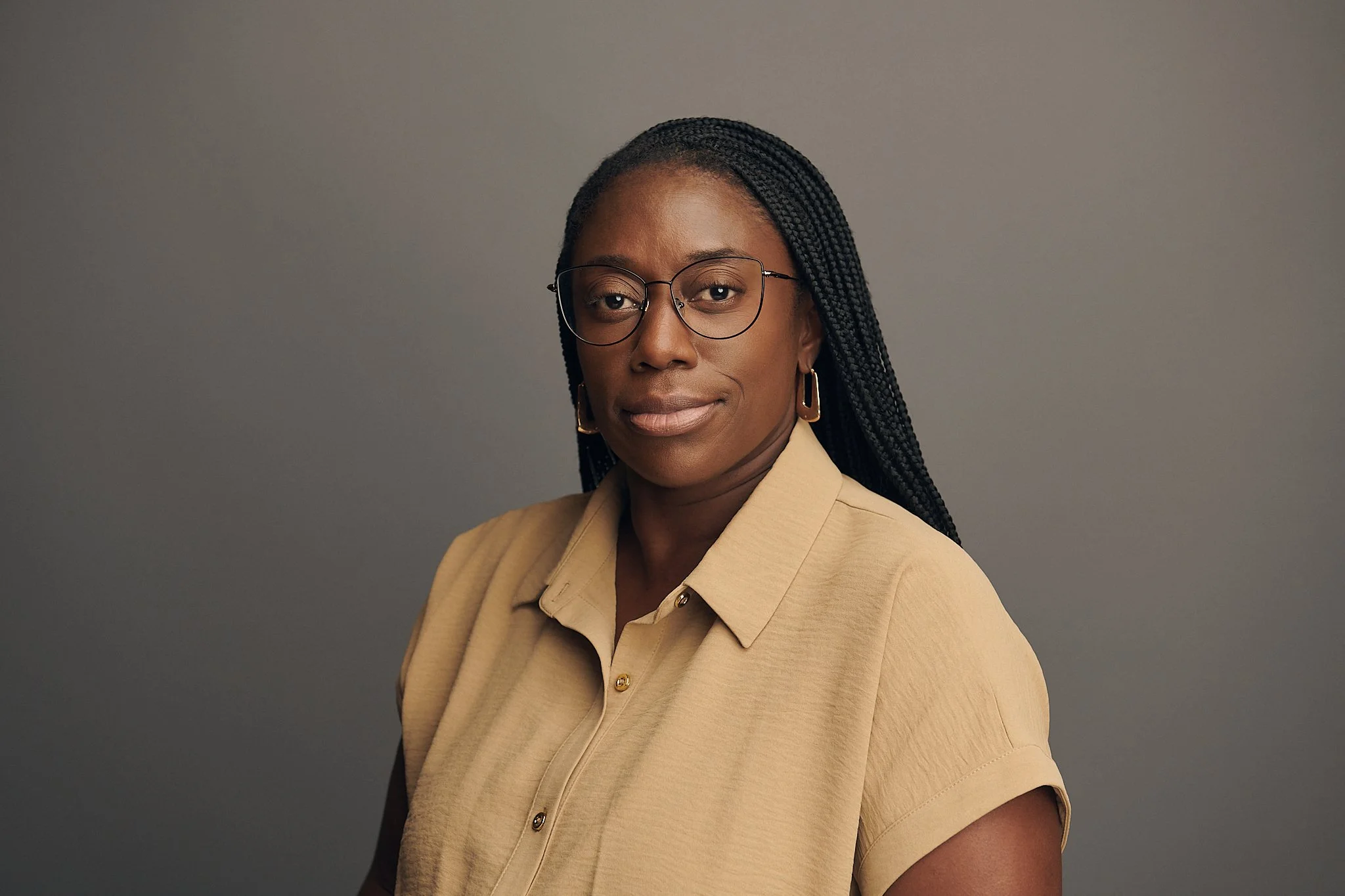 Portrait of a Black woman with long braided hair, wearing glasses, a beige shirt with roll-up sleeves, and gold hoop earrings, against a plain gray background.