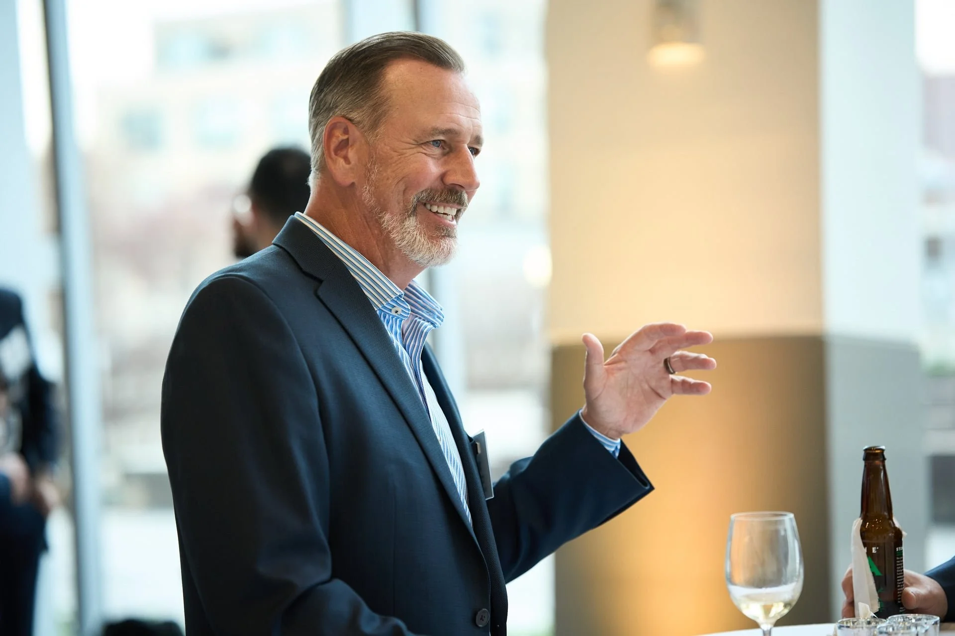 A middle-aged man with short gray hair and a beard, dressed in a dark suit and striped shirt, is smiling and gesturing with his right hand during a social gathering or event.