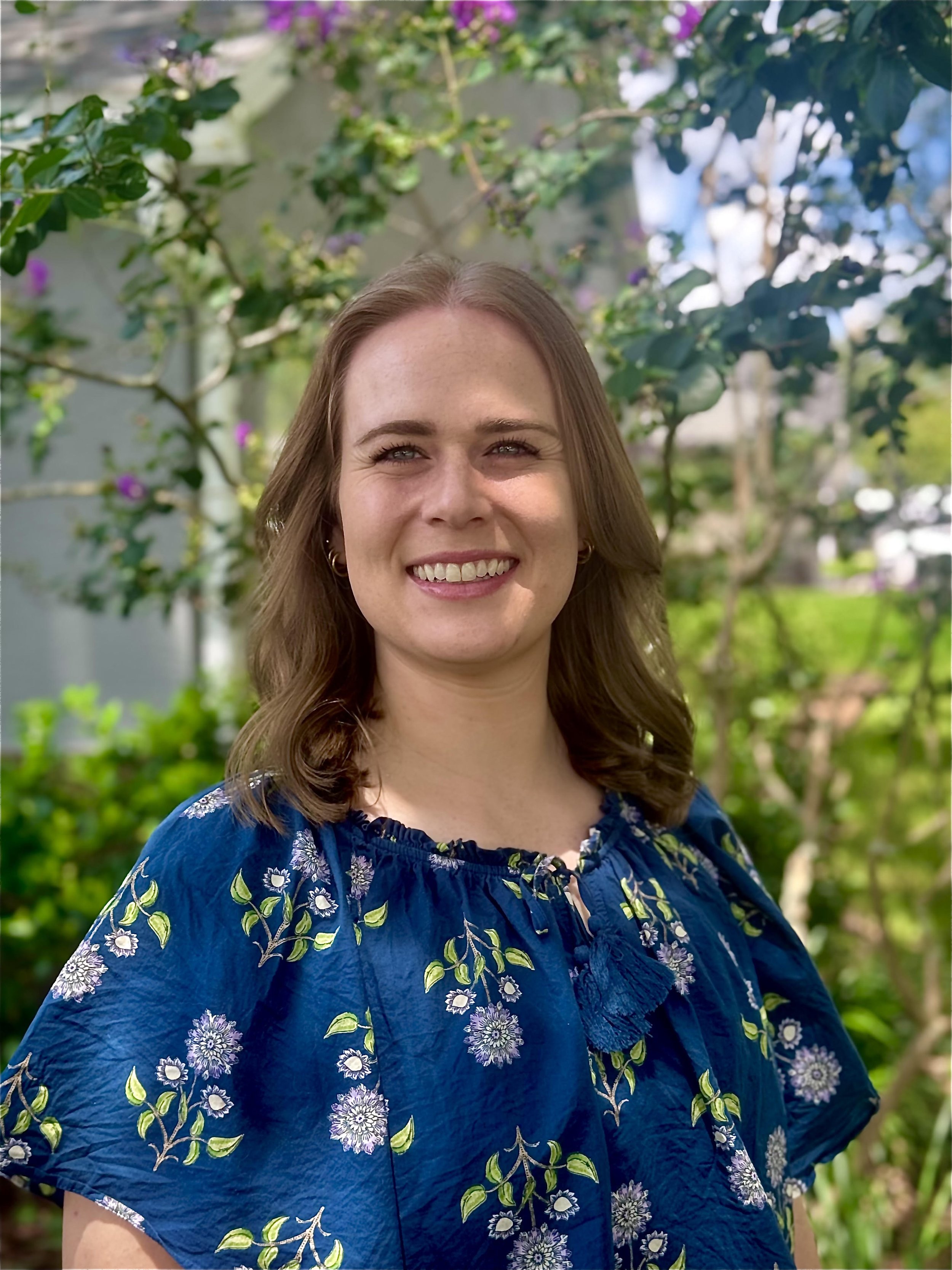 A woman with light brown, wavy hair smiling outdoors, wearing a blue blouse with a floral pattern, surrounded by green trees and purple flowers.