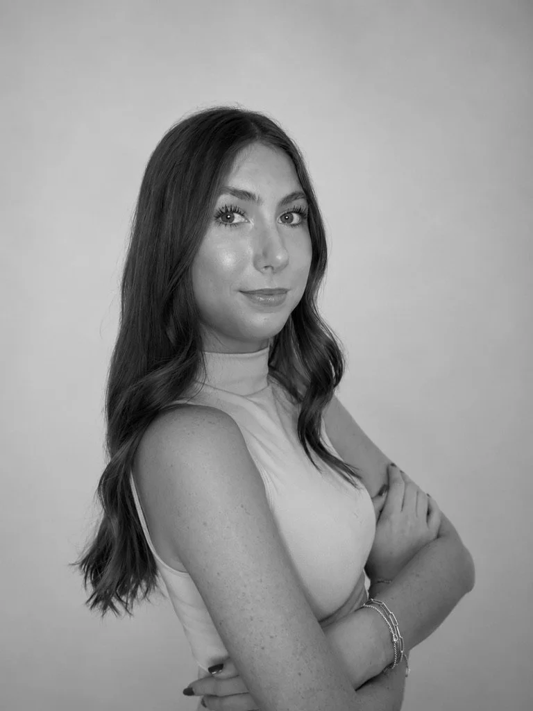 Black and white portrait of a young woman with long wavy hair, wearing a sleeveless top, crossing her arms, posing confidently.