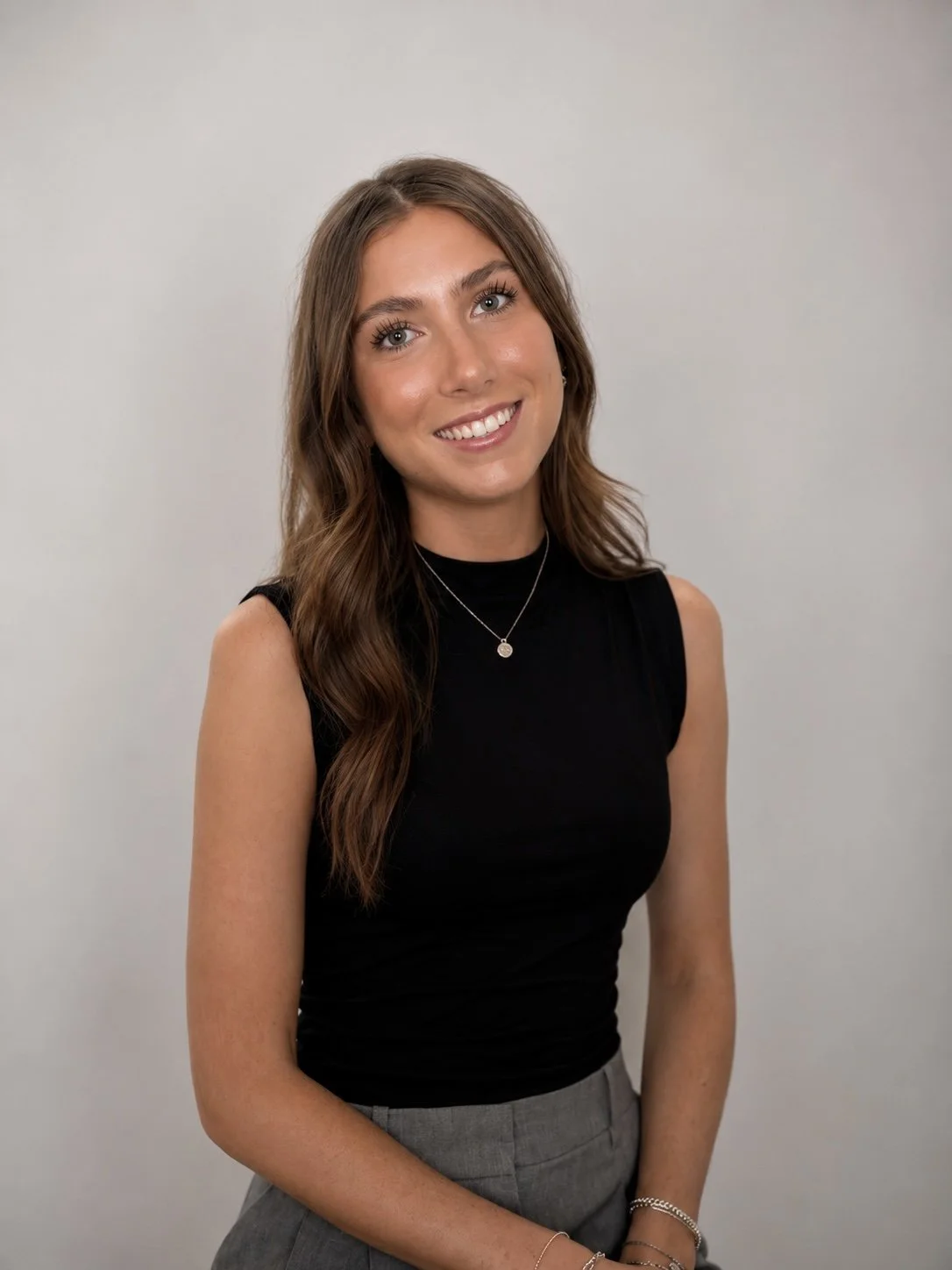 Young woman with long wavy brown hair wearing a black sleeveless top, gray pants, and layered jewelry, smiling against a plain gray background.