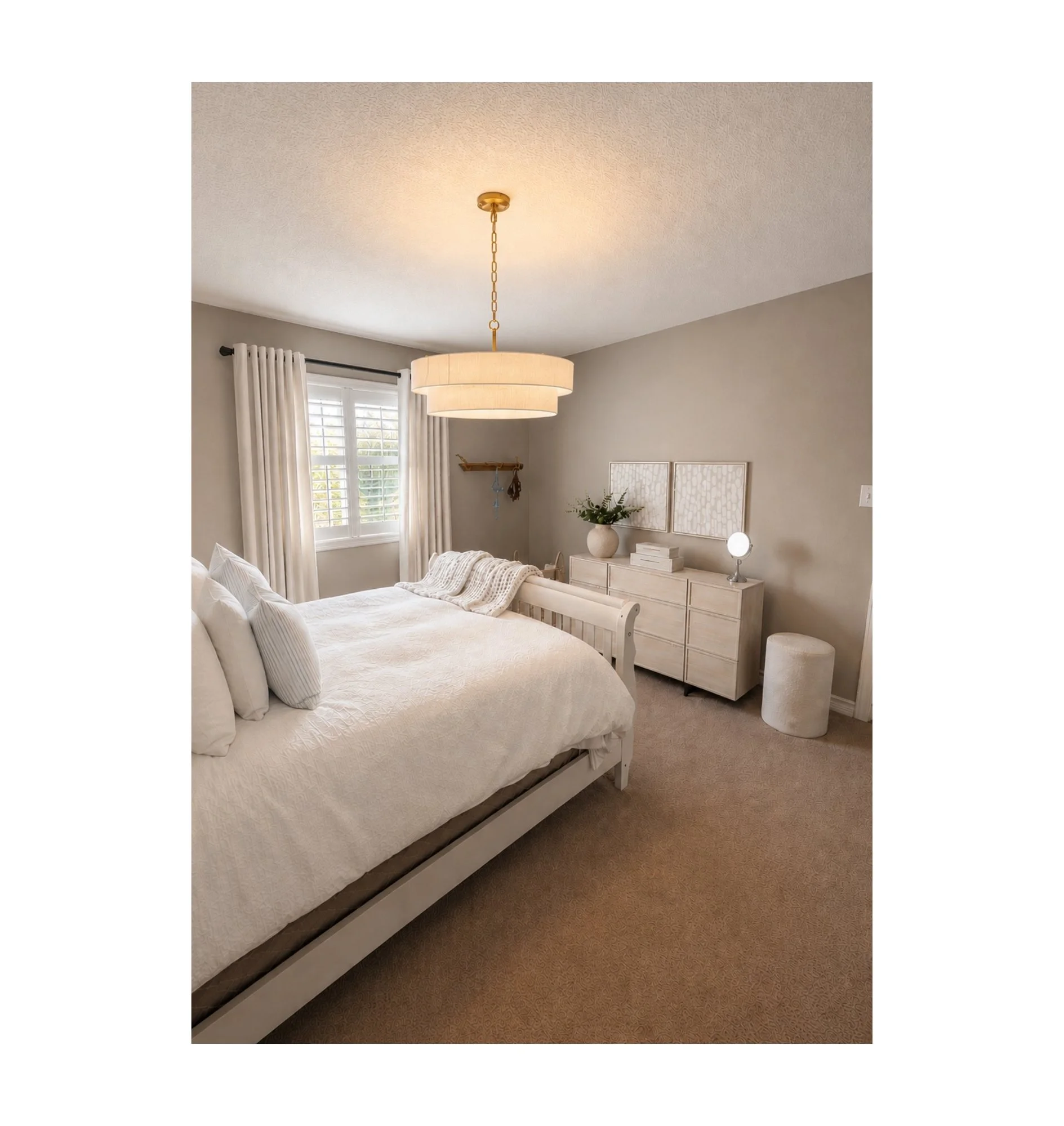 A bedroom with beige carpet, white bed with pillows and blanket, window with white curtains, desk with a mirror and small stool, and ceiling light fixture.