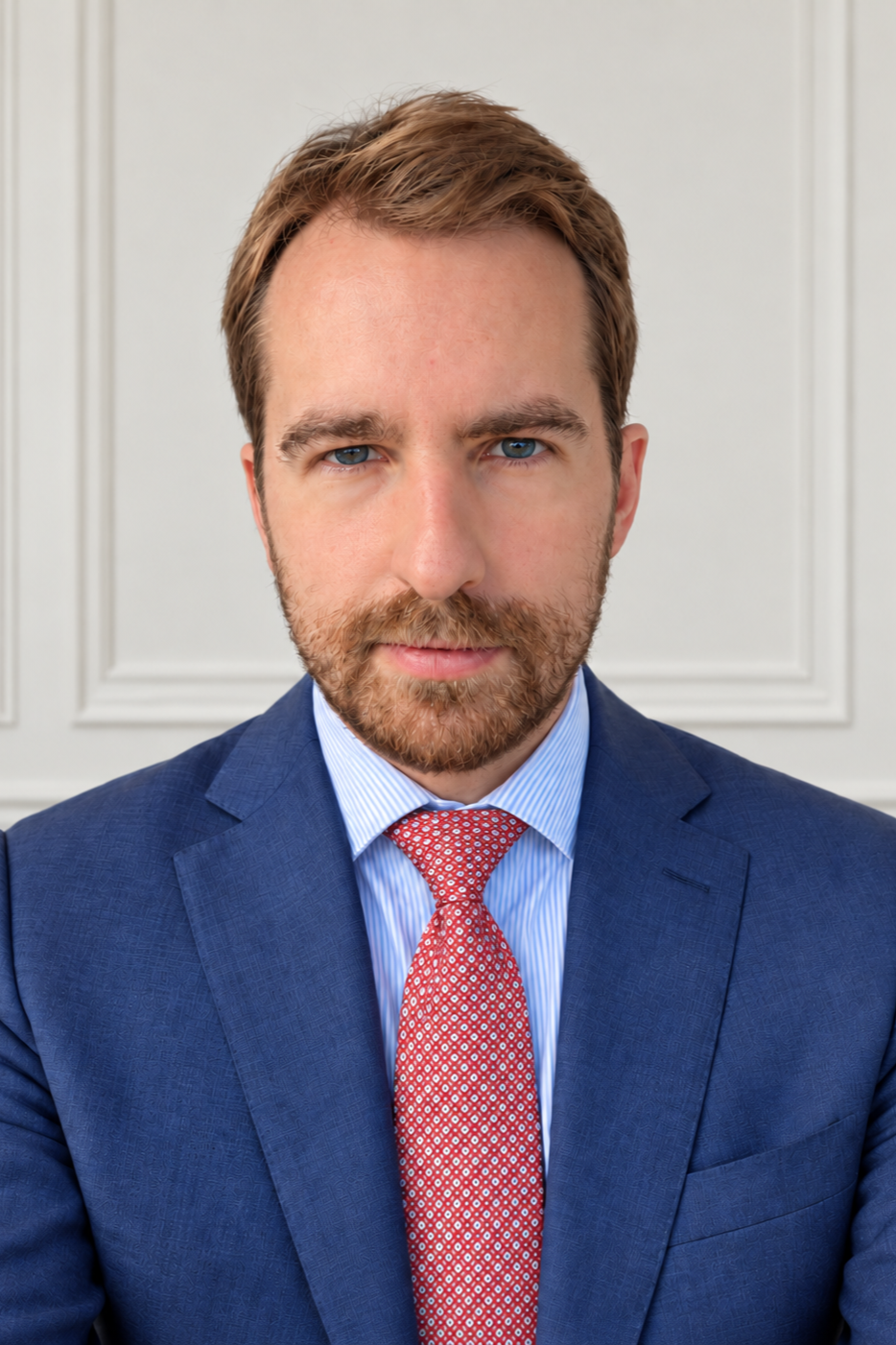 A man with short brown hair, blue eyes, and a beard in a blue suit and red patterned tie, standing in front of a white wall with decorative molding.