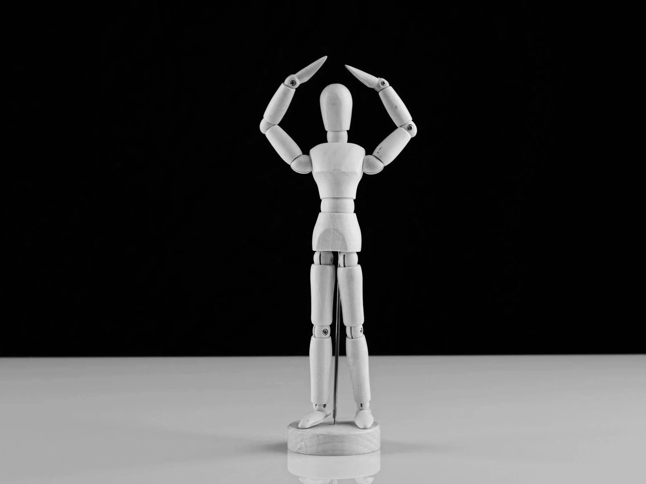 A white wooden mannequin figure standing on a small round base, holding its hands above its head. The background is black and the figure is positioned on a flat surface. Good posture