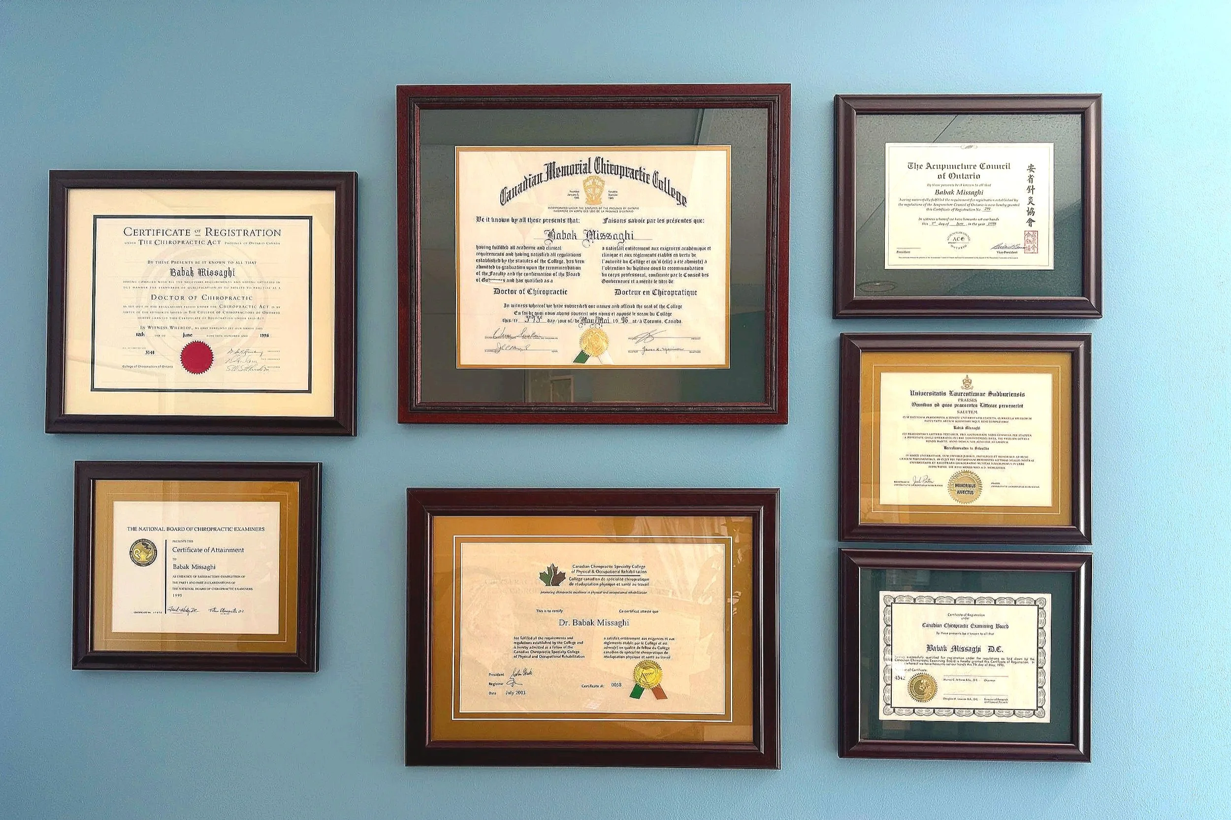 Wall with multiple framed certificates and diplomas, including degrees and professional achievement awards, arranged in a grid pattern on a light blue wall. Diplomas Dr. Babak Missaghi, Vitual consultation