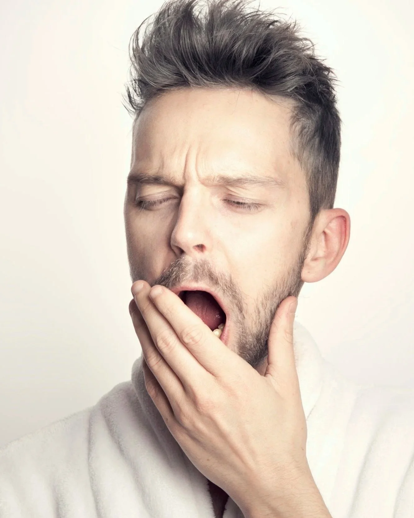 A man with short brown hair and a beard yawning with eyes closed and mouth open, hand covering part of his chin, against a plain light background. TMJ