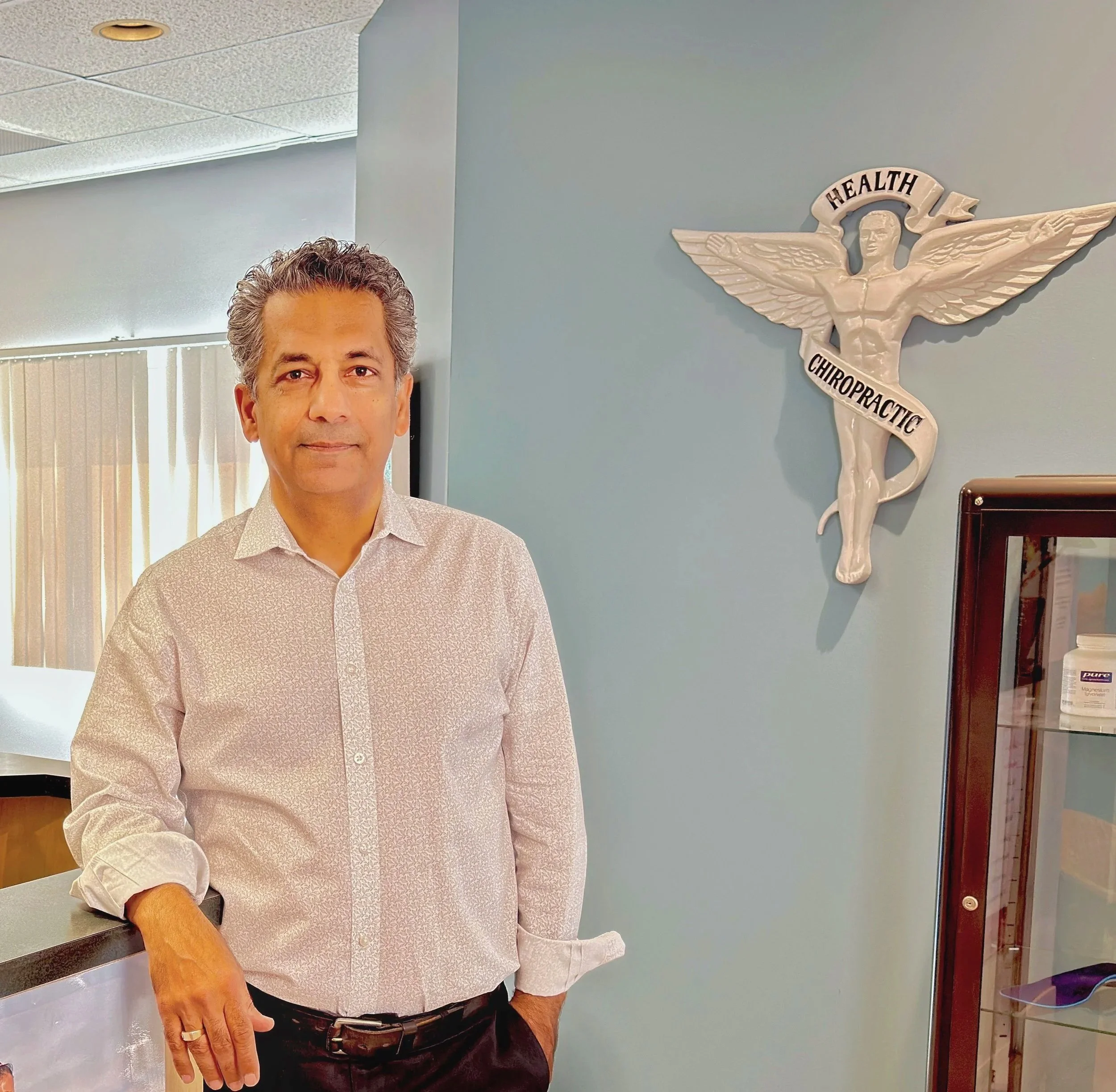 A man with dark hair and a light patterned shirt standing in an office or clinic, with a blue wall behind him. On the wall is a white emblem of a female figure with wings and the words 'Health Chiropractic'. Dr. Babak Missaghi