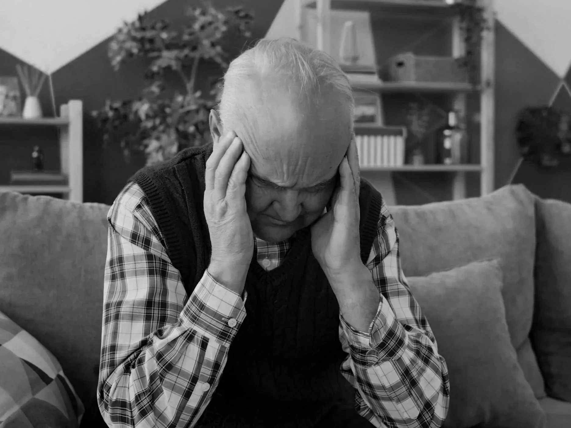 Older man with white hair, wearing checkered shirt and dark vest, sitting on a couch with hands on his head, in a living room with shelves and plants in the background. Vertigo