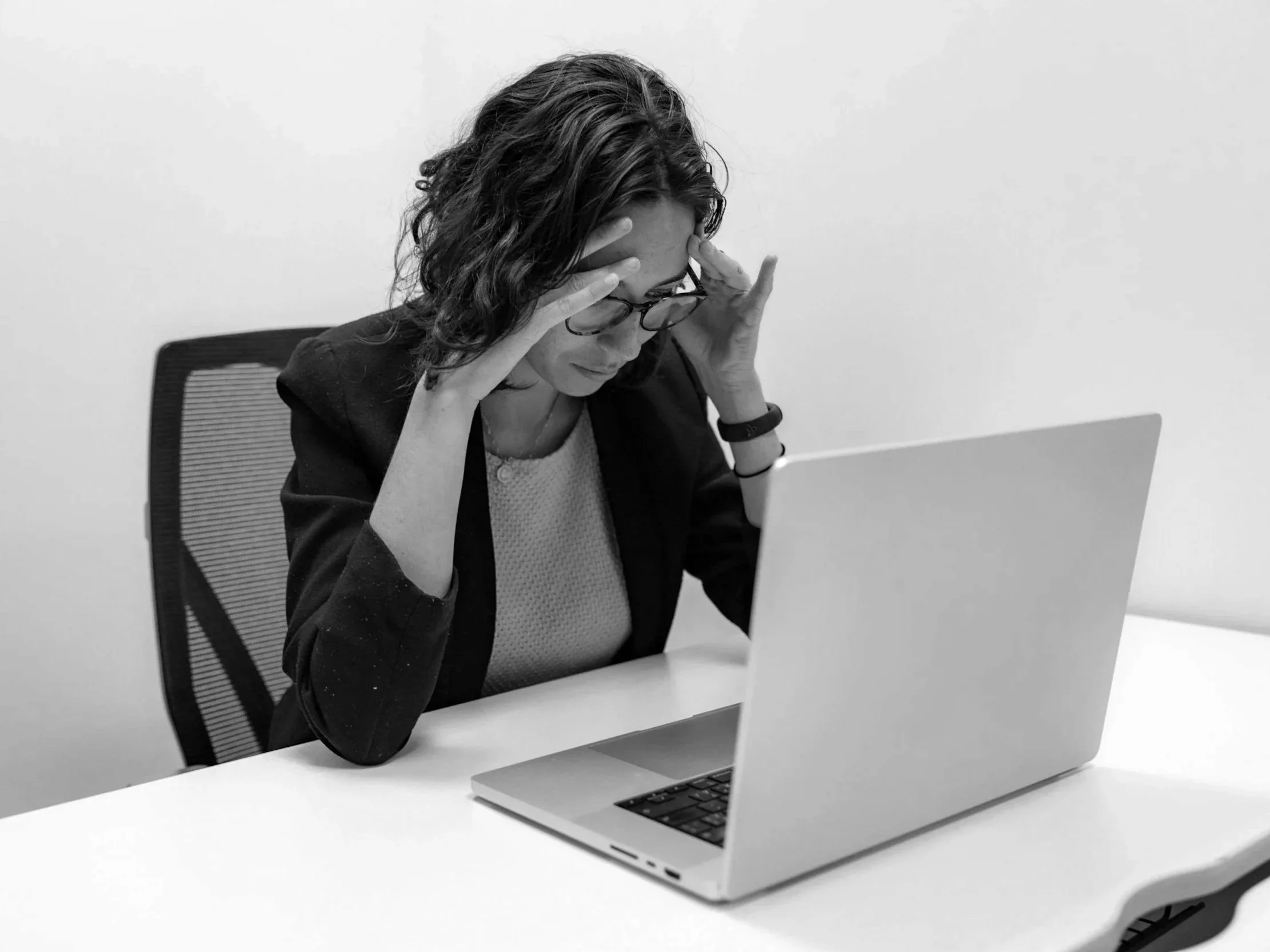 A woman with glasses and curly hair is sitting at a desk with her hands on her head, looking at a laptop. The setting appears to be an office with a plain white wall in the background. Headache