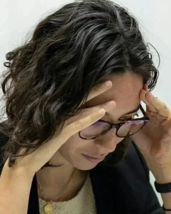A woman with short, curly dark hair and glasses holding her head with both hands, appearing stressed or worried. Headache